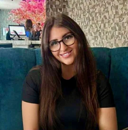 A young woman with long brown hair and glasses smiling at the camera, sitting on a teal-colored booth in a cafe with pink trees and a patterned wall in the background.