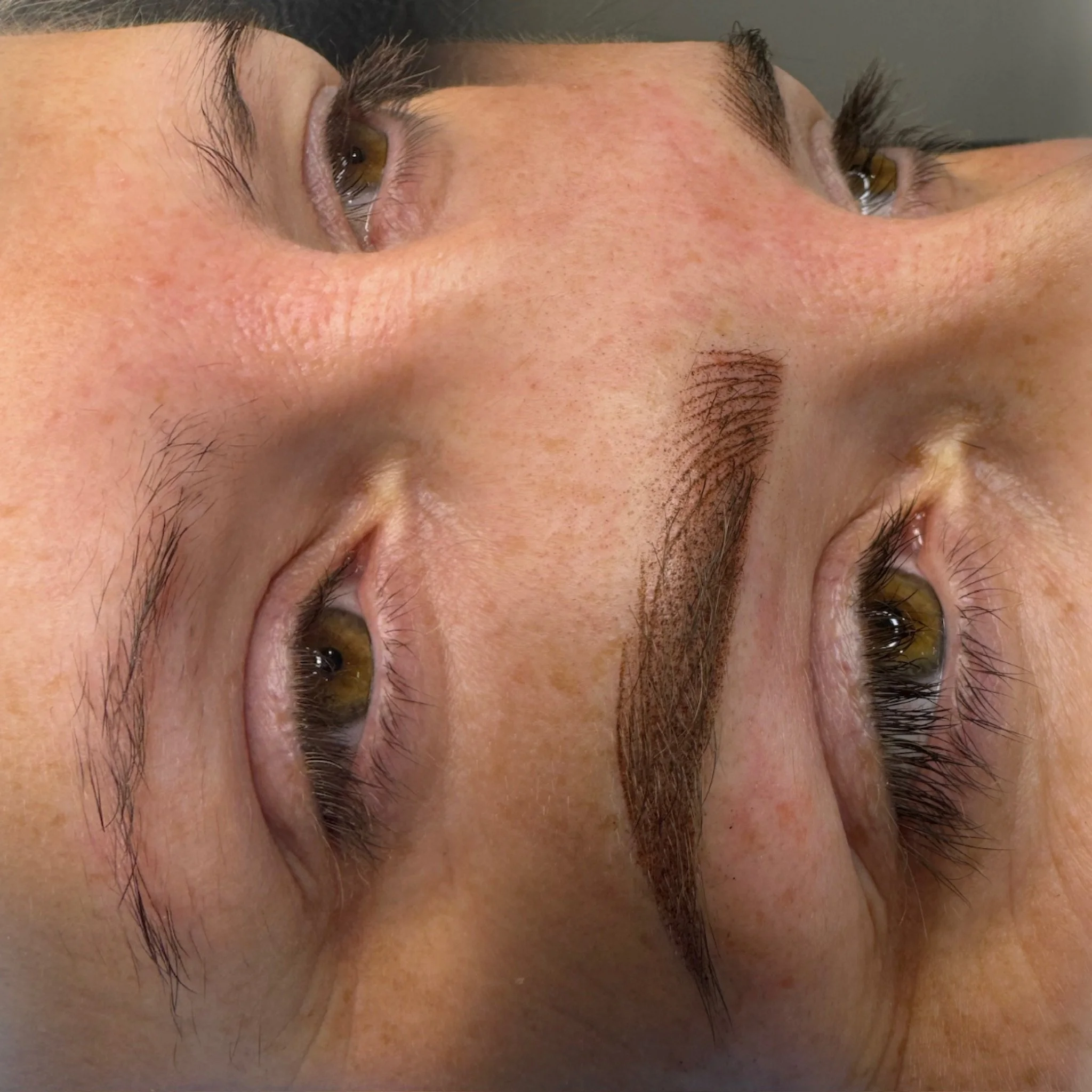 Close-up photo of a person's face with a focus on their brown eyebrows, light skin with freckles, and dark eyes, lying down with a neutral background.