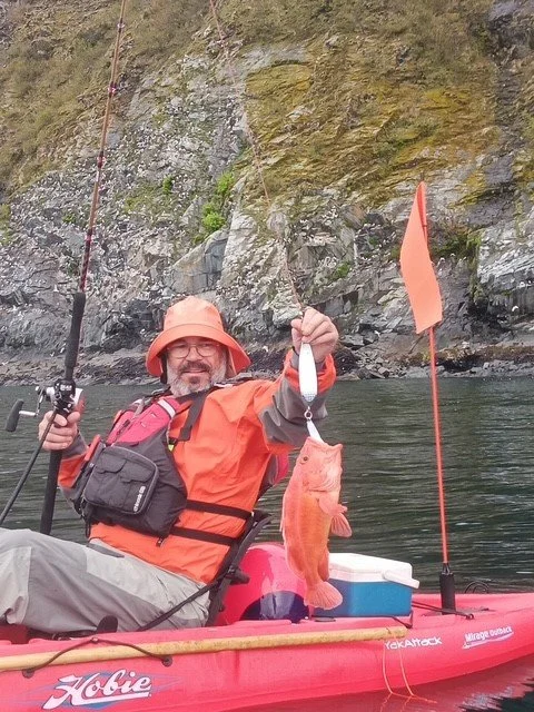 Man in orange jacket and hat sitting in a kayak holding a fish he caught, with a rocky shoreline in the background.