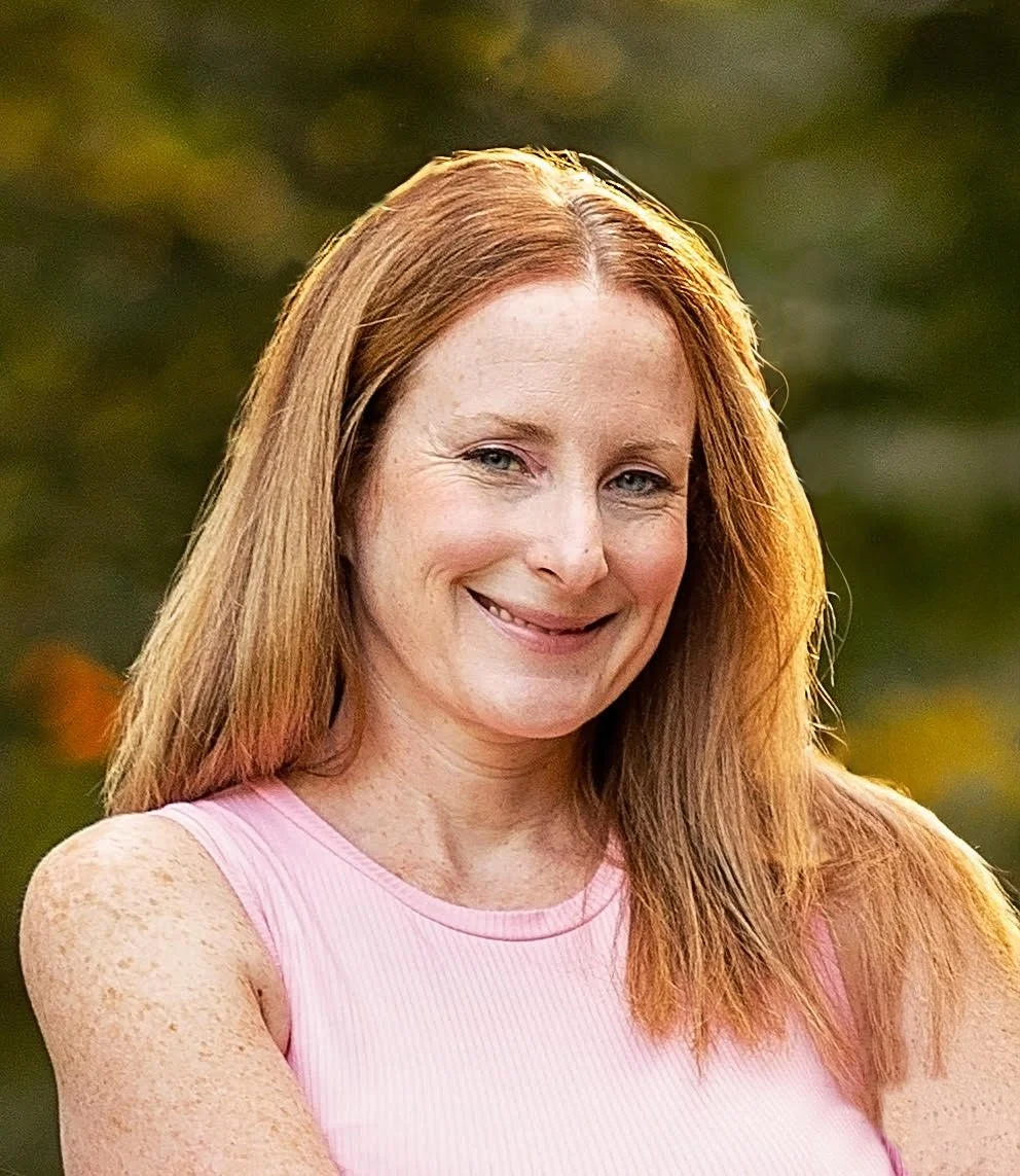A woman with long red hair smiling outdoors, wearing a pink sleeveless top, with a blurred nature background.