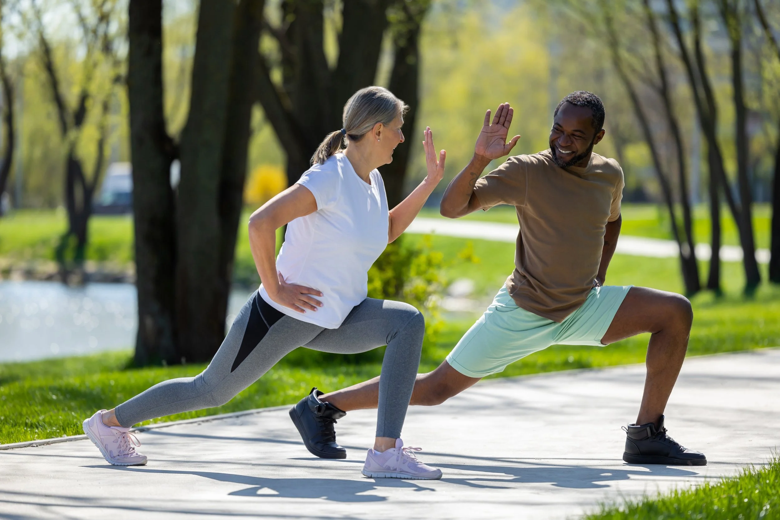 A woman and a man exercising outdoors in a park, doing lunges and high-fives.