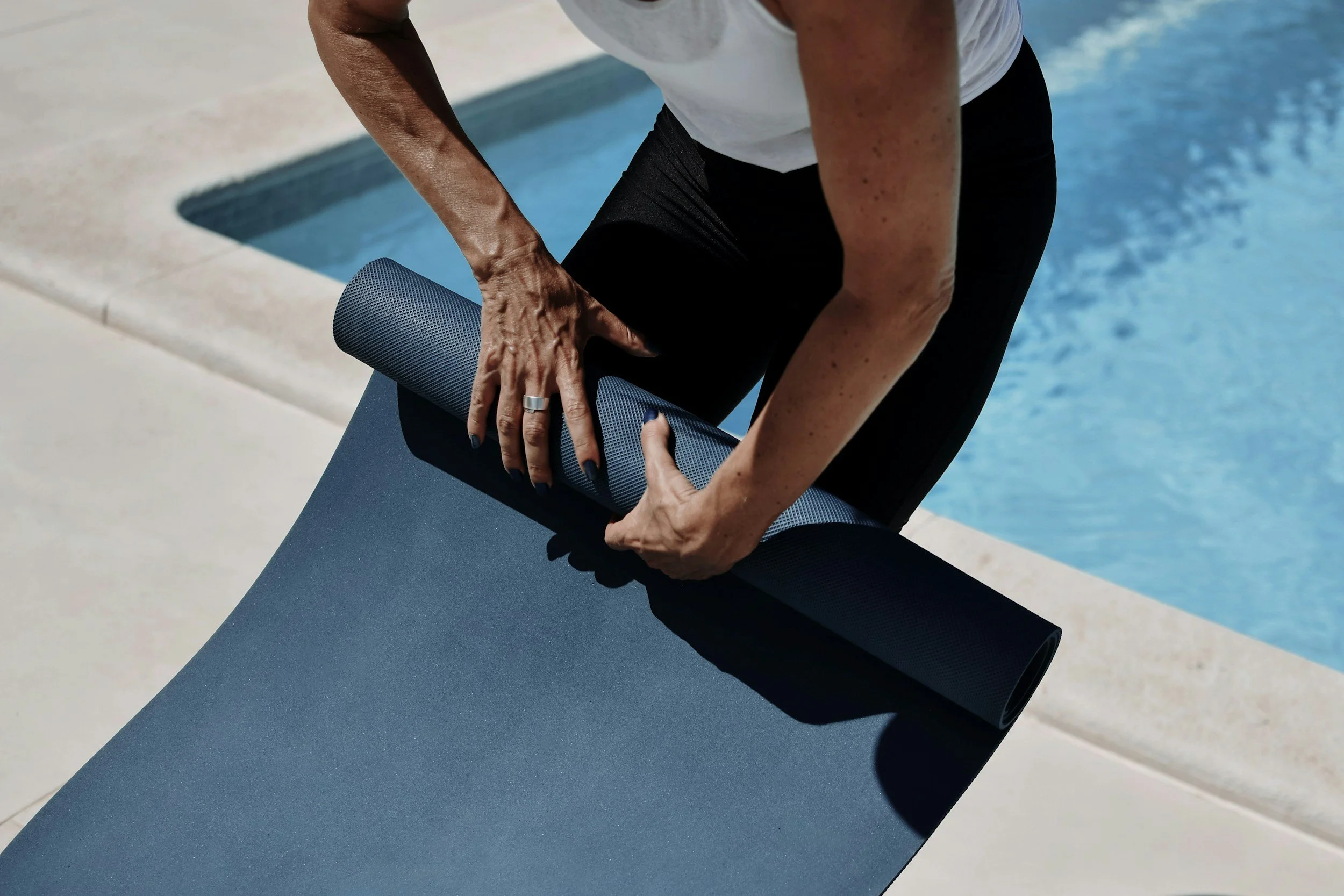 Person rolling up a black yoga mat beside a swimming pool