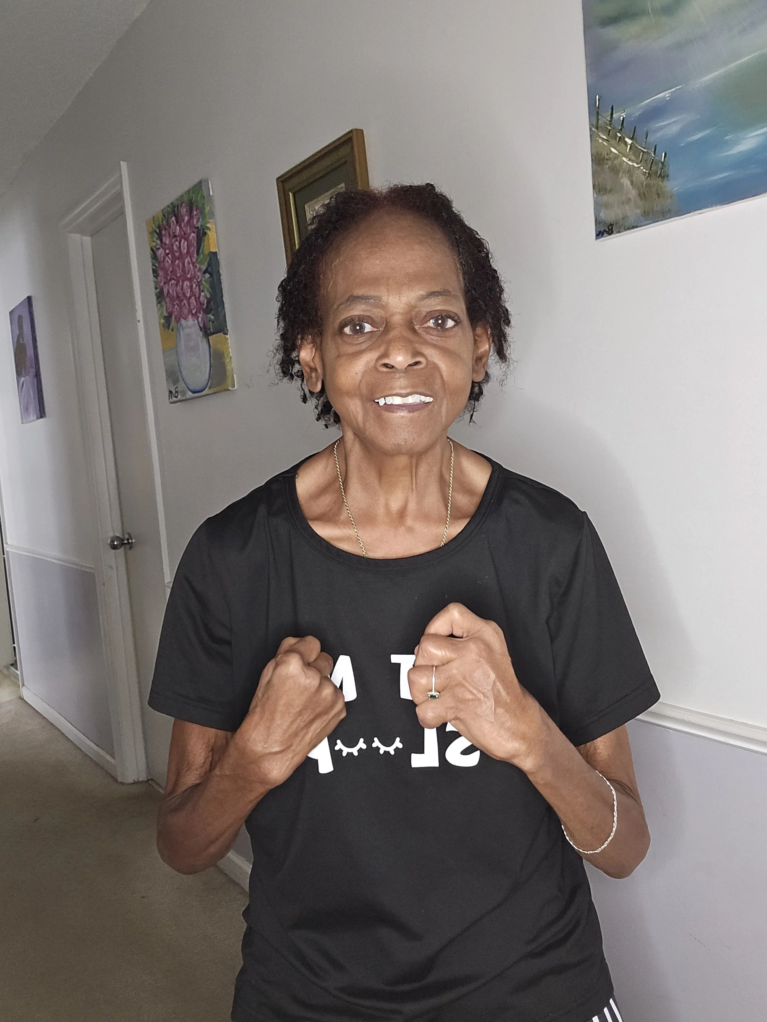 An elderly woman with short, curly hair standing indoors, wearing a black t-shirt and holding her fists up in a fighting stance.