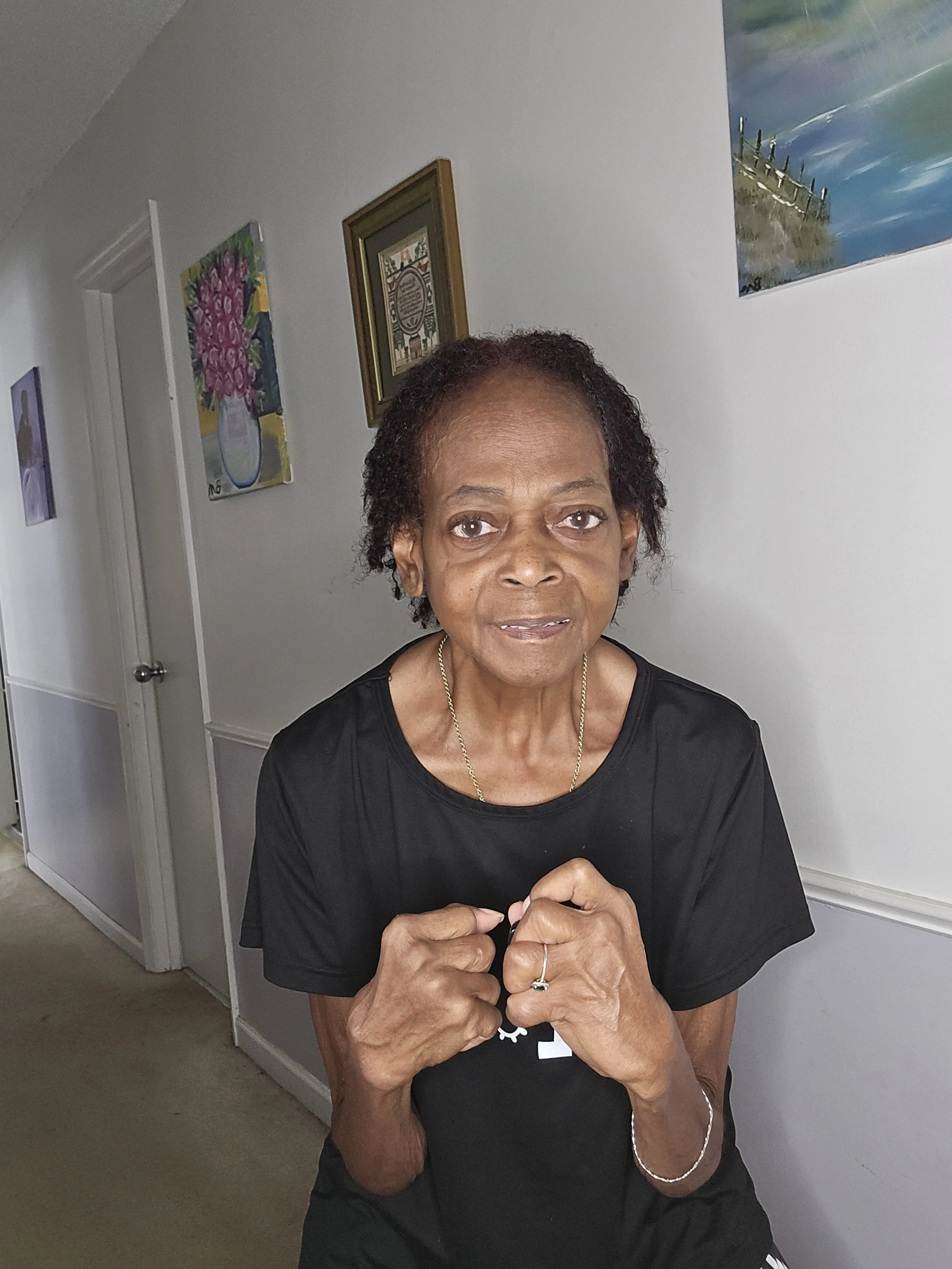 Portrait of an elderly woman with short curly hair, wearing a black shirt, standing indoors with her hands clasped in front of her, background includes framed artwork on a white wall.