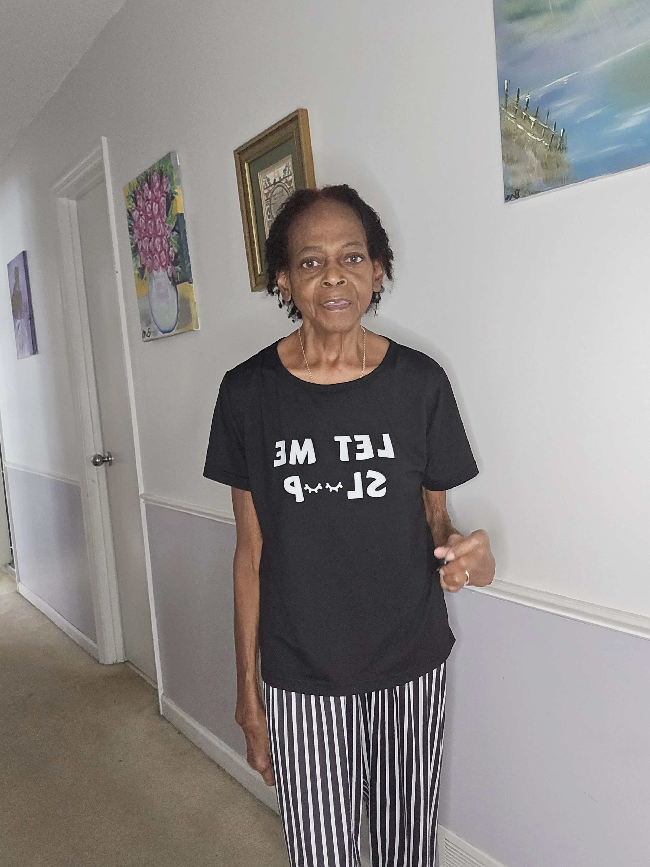 An elderly woman standing indoors, wearing a black T-shirt with white text that says 'Let Me Sleep' and black-and-white striped pants. The background shows framed artwork on a light-colored wall.