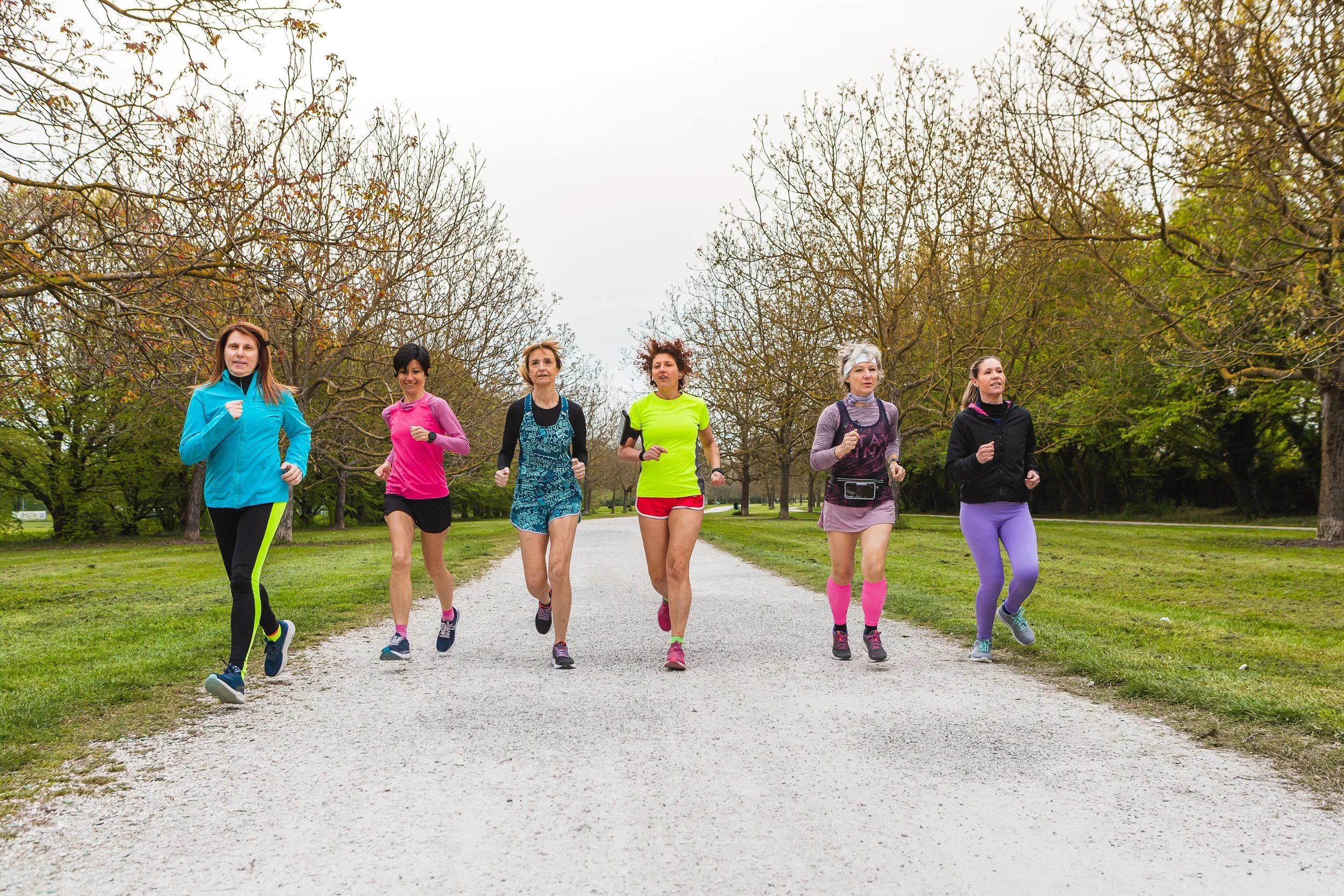 Six women running together on a park trail, surrounded by trees with some leaves, wearing colorful athletic clothing.