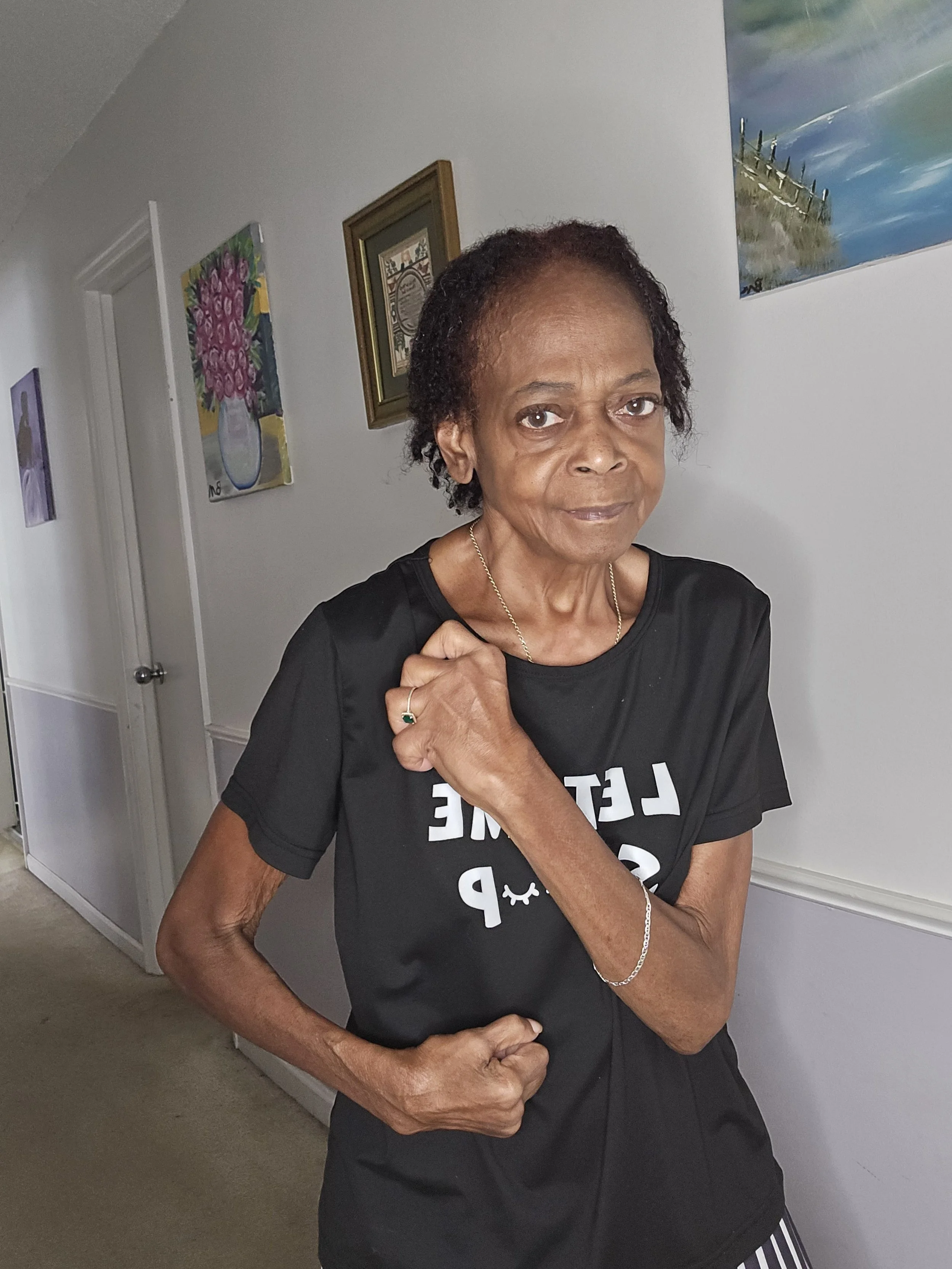 An elderly woman with short curly hair standing indoors, wearing a black T-shirt with white text, and accessorized with a necklace, a bracelet, and a ring. She appears to be in a room with artwork on the wall.