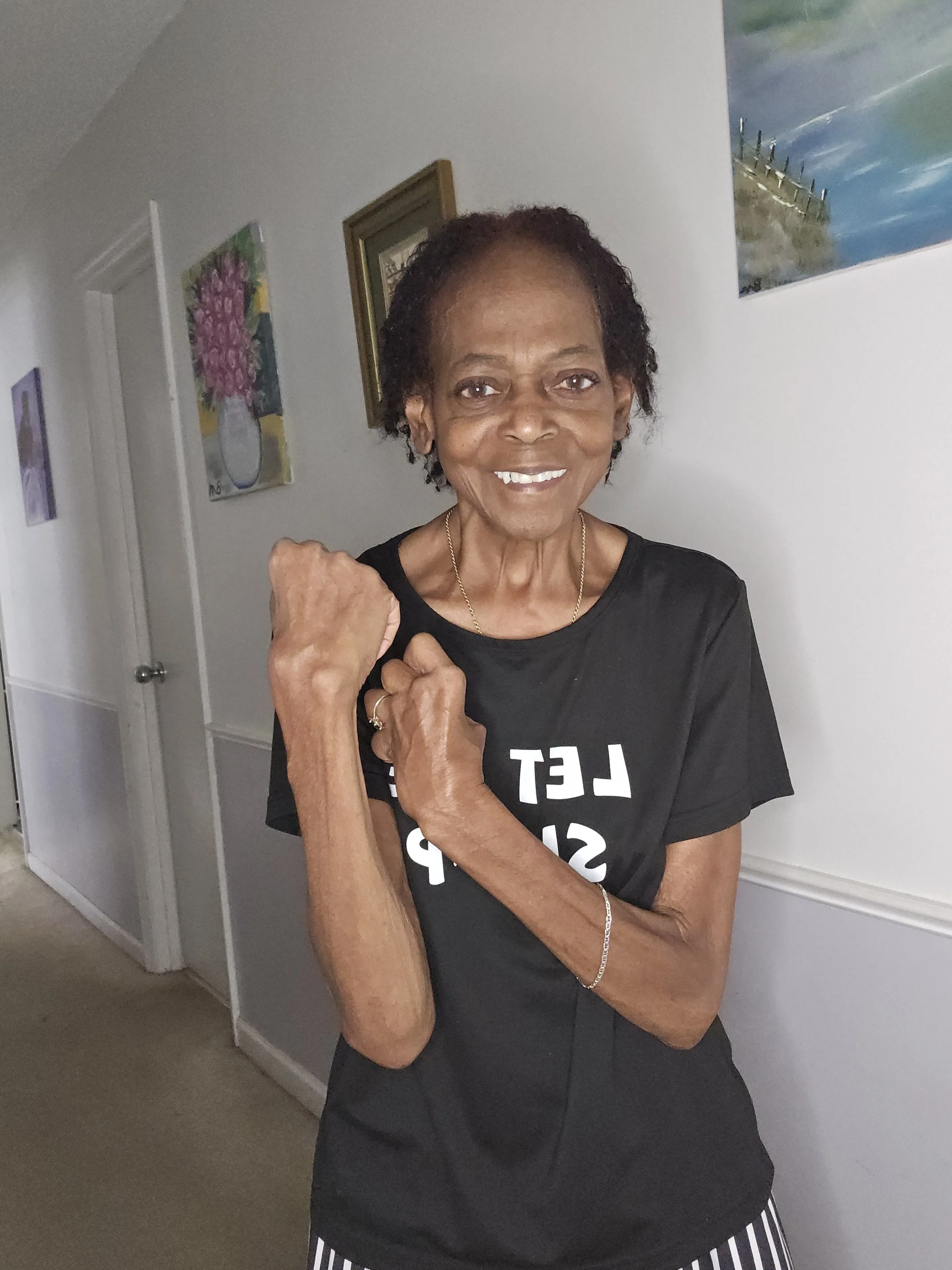 An elderly woman smiling, wearing a black T-shirt with white text and jewelry, standing indoors in front of a white wall with framed artwork.
