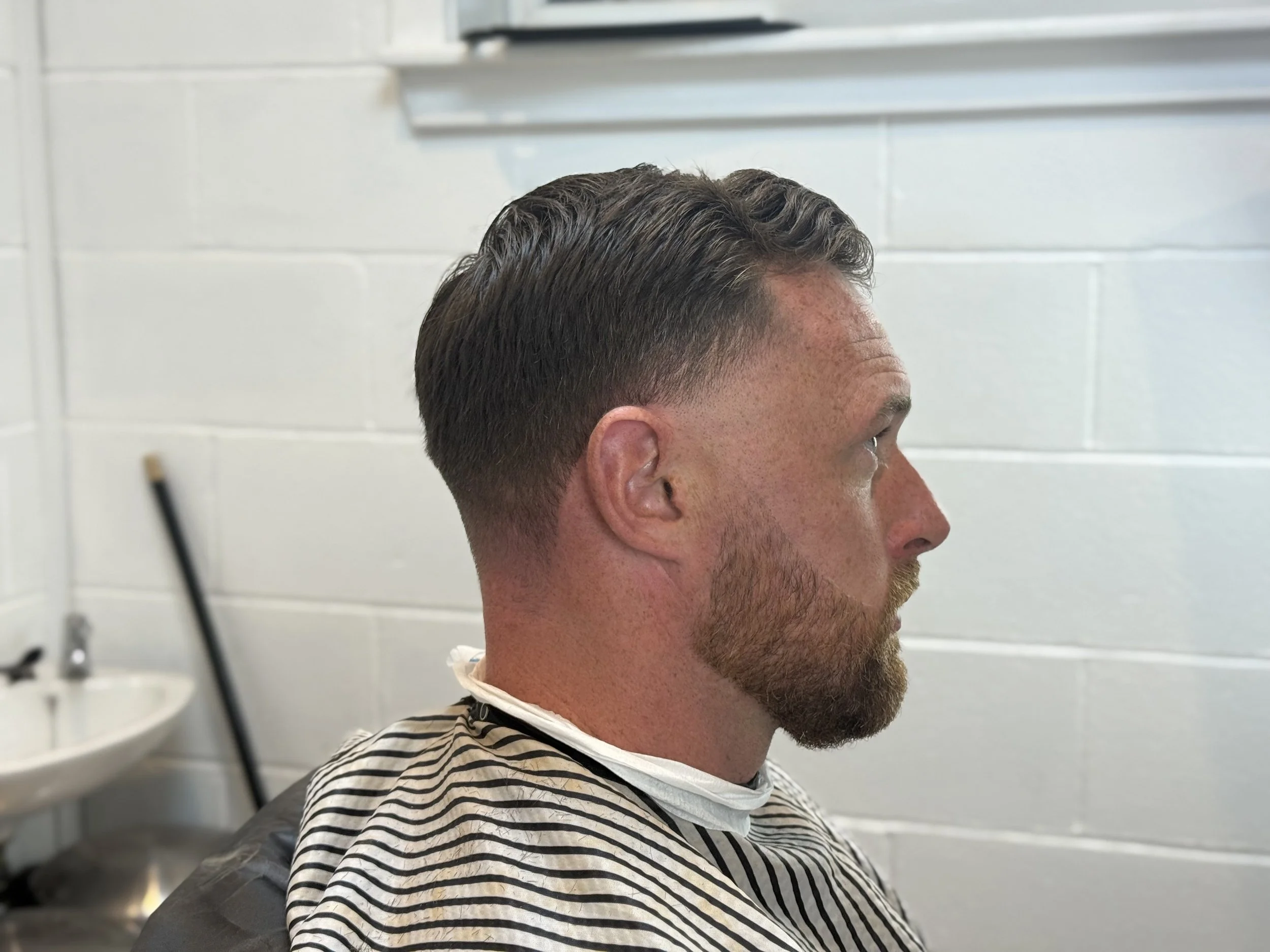Side profile of a man with a fresh haircut, beard, and mustache, wearing a striped barber cape, seated in a barber shop. The background features a white tiled wall, a sink, and a broom.