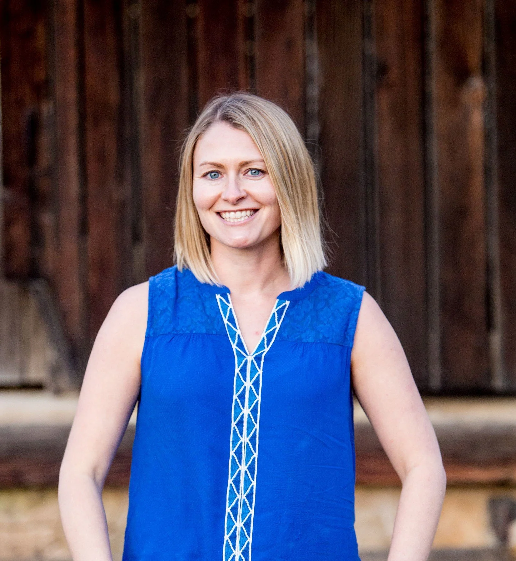 A woman with shoulder-length blonde hair, blue eyes, smiling, wearing a sleeveless blue dress with white decorative stitching, outdoors in front of a wooden fence.