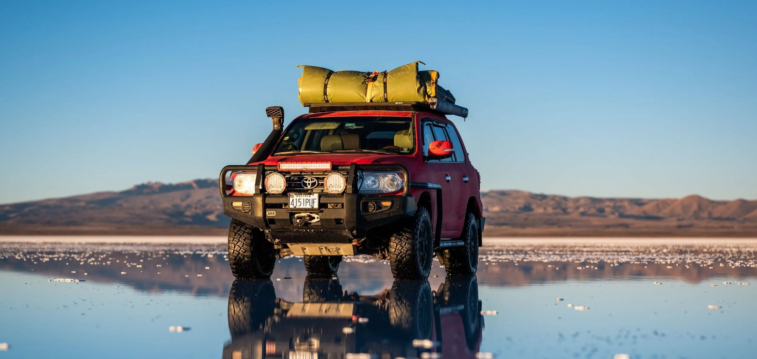 Vehículo todoterreno rojo con equipo de camping en el techo, en un paisaje desértico con agua reflectante y montañas al fondo.