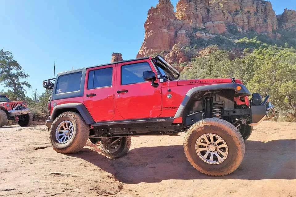 Jeep Rubicon rojo en un terreno arenoso con montañas y árboles verdes en el fondo.