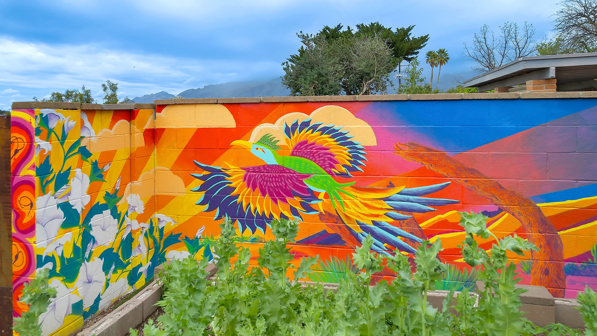 Colorful mural of a bird with vibrant feathers on a brick wall, with greenery in the foreground and partly cloudy sky in the background.