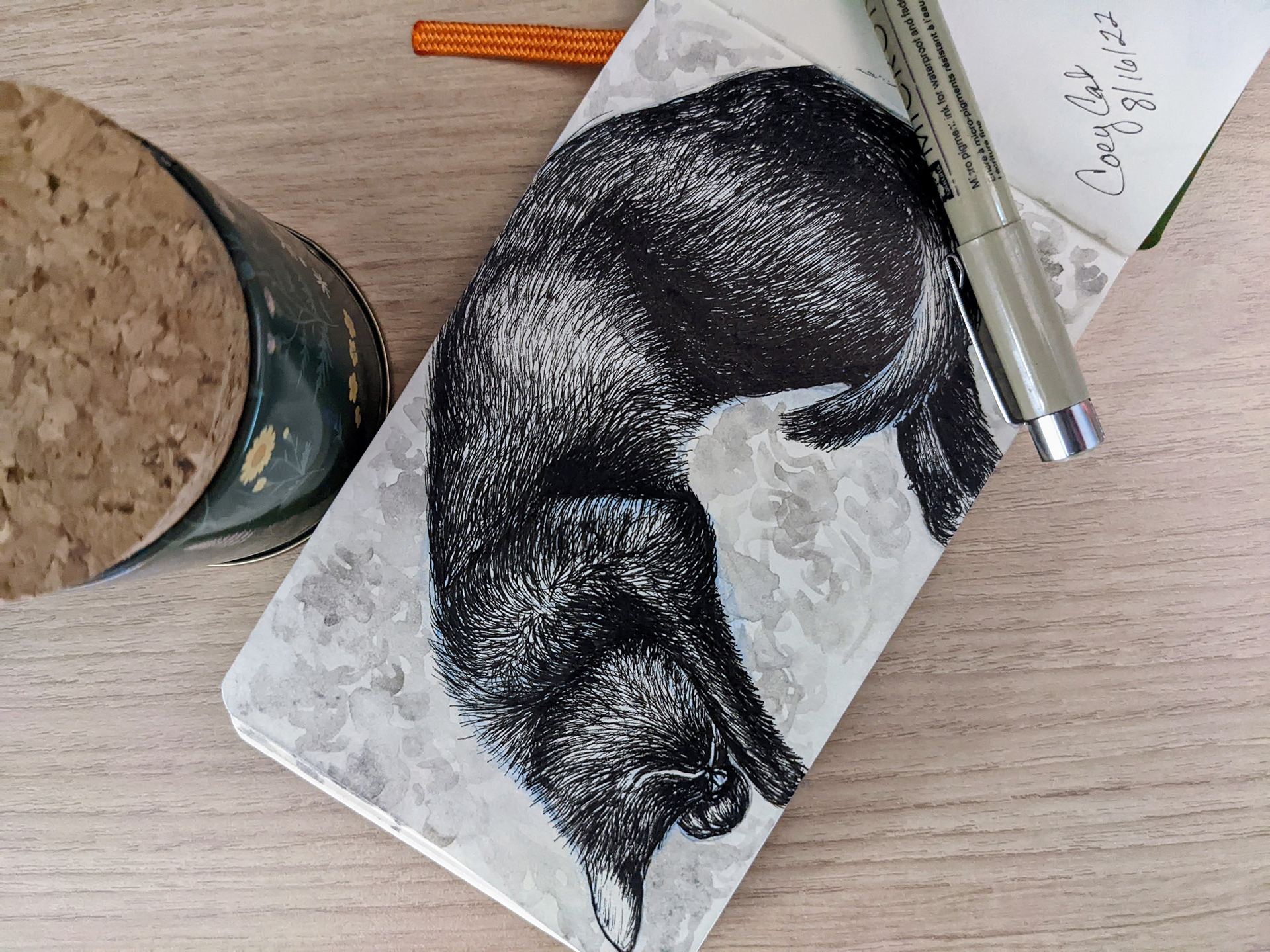 An ink drawing featuring a detailed black ink drawing of a sleeping cat on a floral patterned background, accompanied by a silver pen, a handwritten note, and a jar of tea or coffee on a wooden surface.