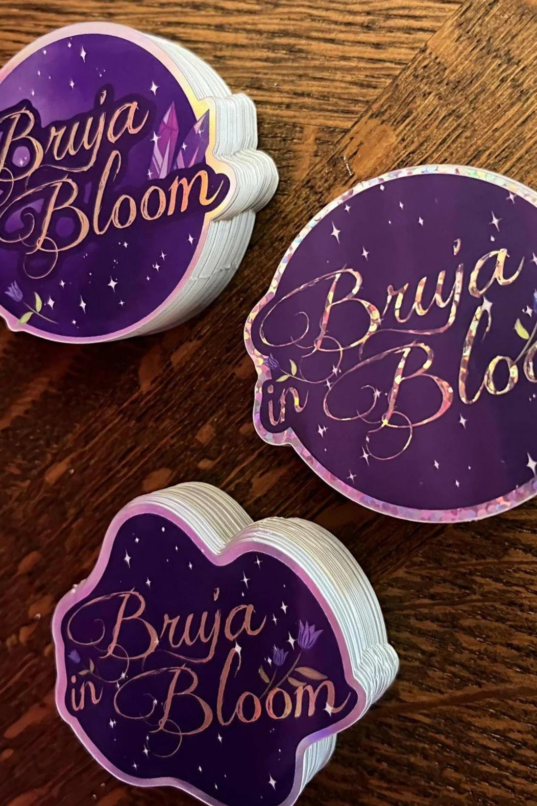 Stacks of round stickers with a purple background, stars, and flowers, reading 'Bruja in Bloom' in elegant script, on a wooden surface.