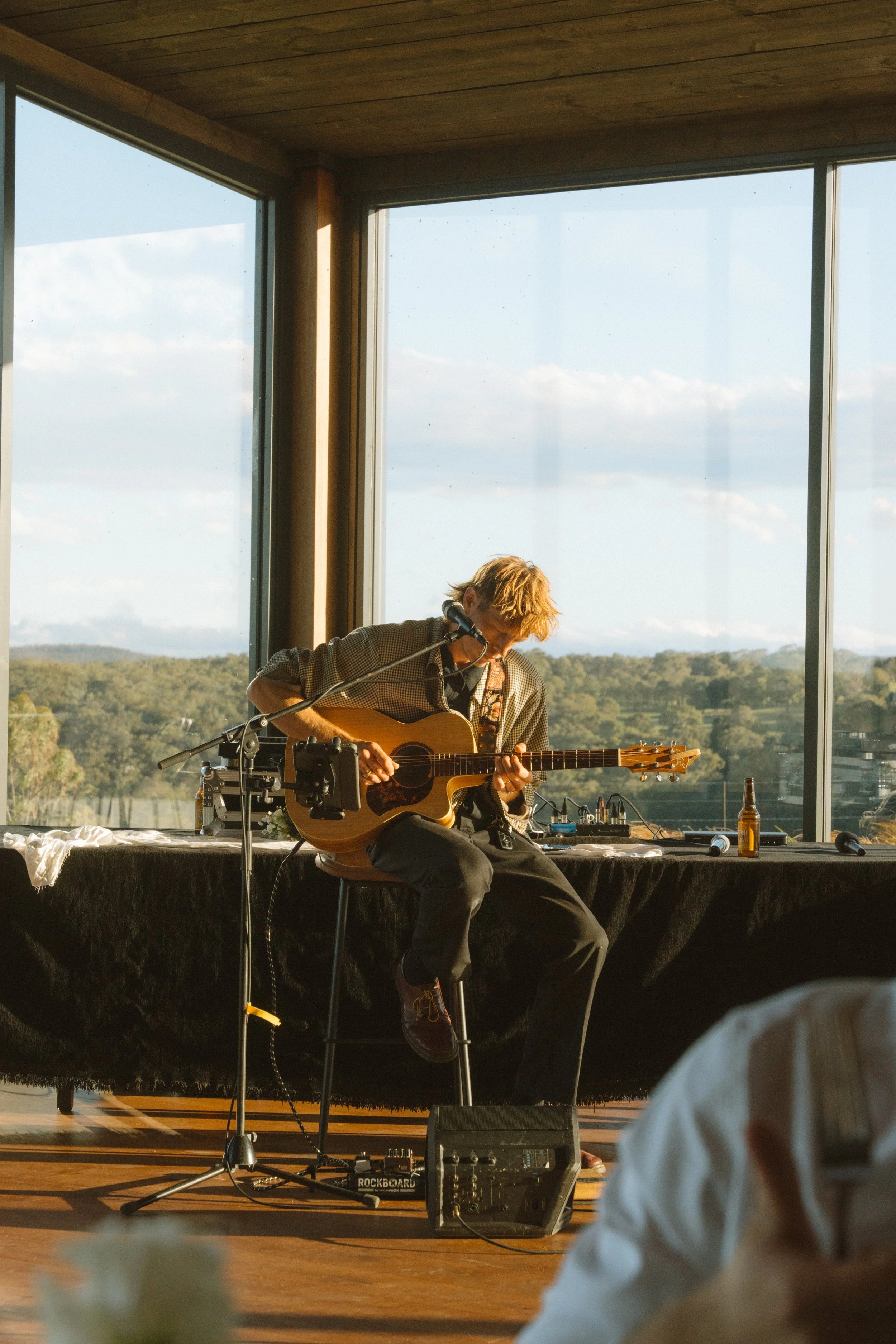 A man playing an acoustic guitar and singing into a microphone during a live performance by a large window with a scenic view of trees and a blue sky.