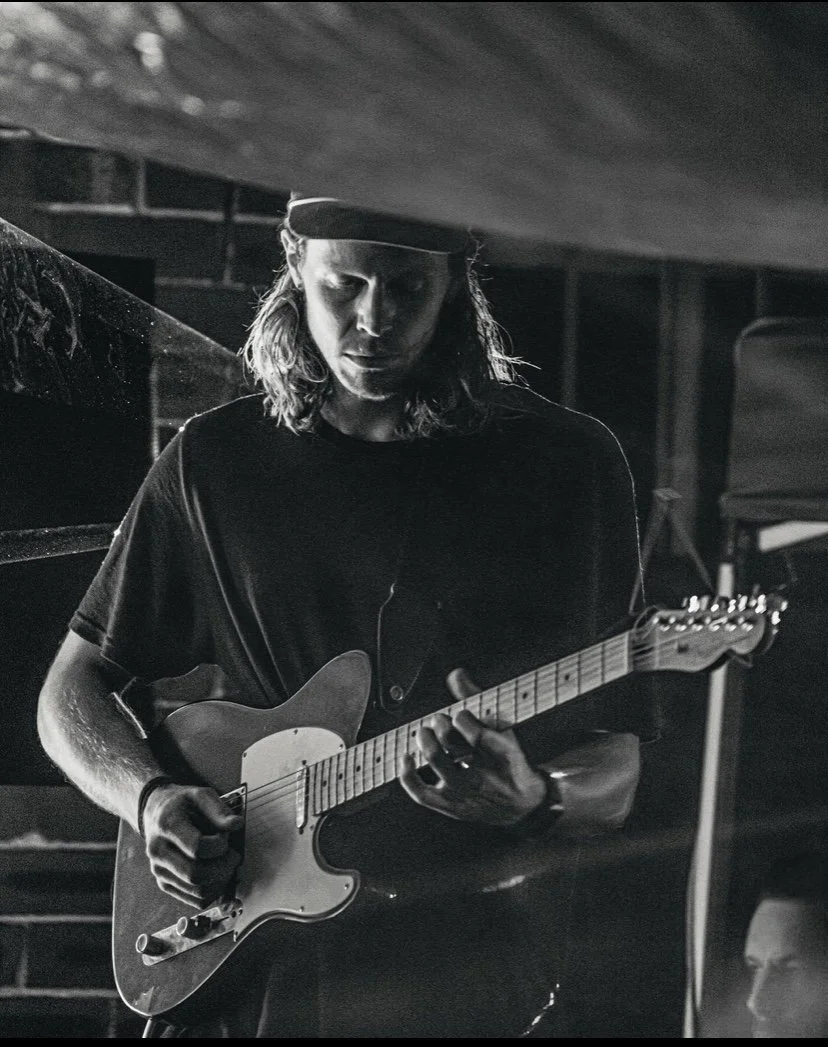 A young man with long hair wearing a baseball cap plays an electric guitar in a dimly lit setting, possibly a stage or studio.