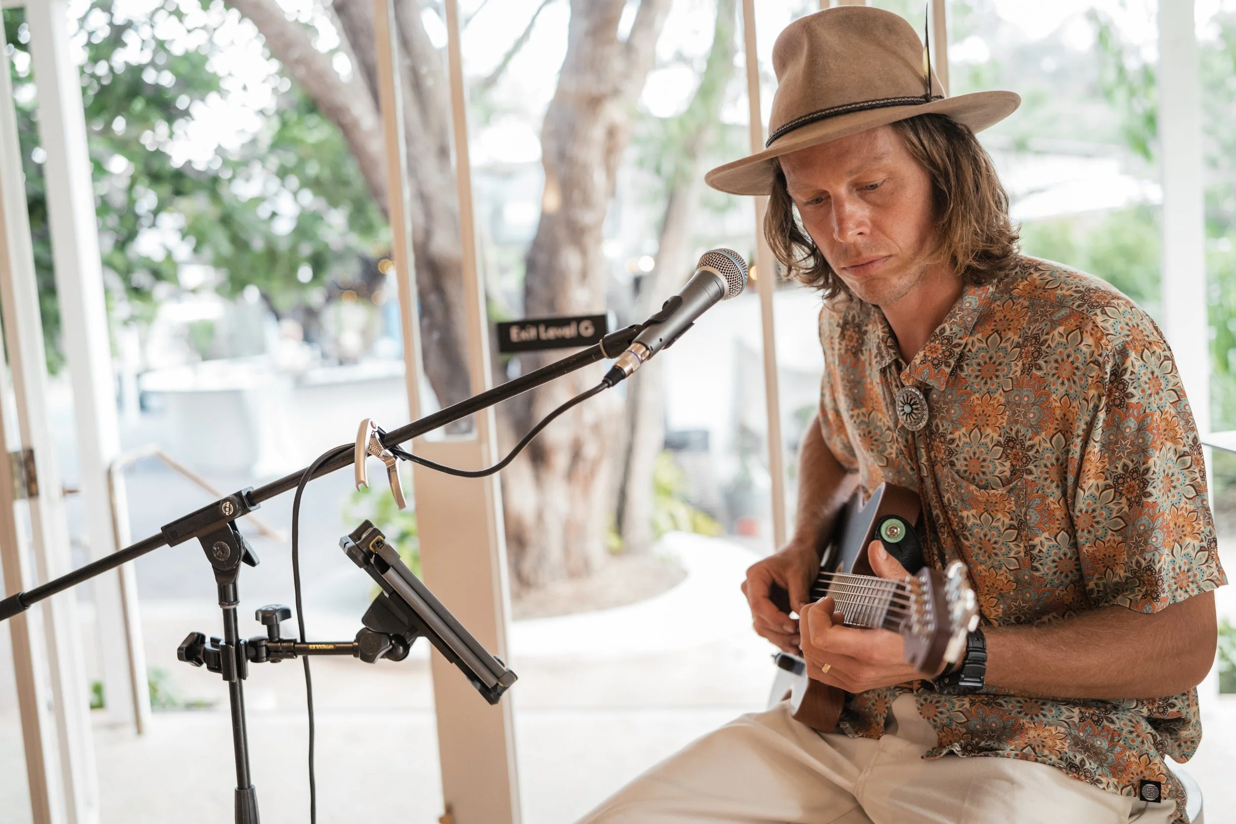 A man wearing a tan hat and a floral shirt playing an acoustic guitar while sitting in front of a microphone.