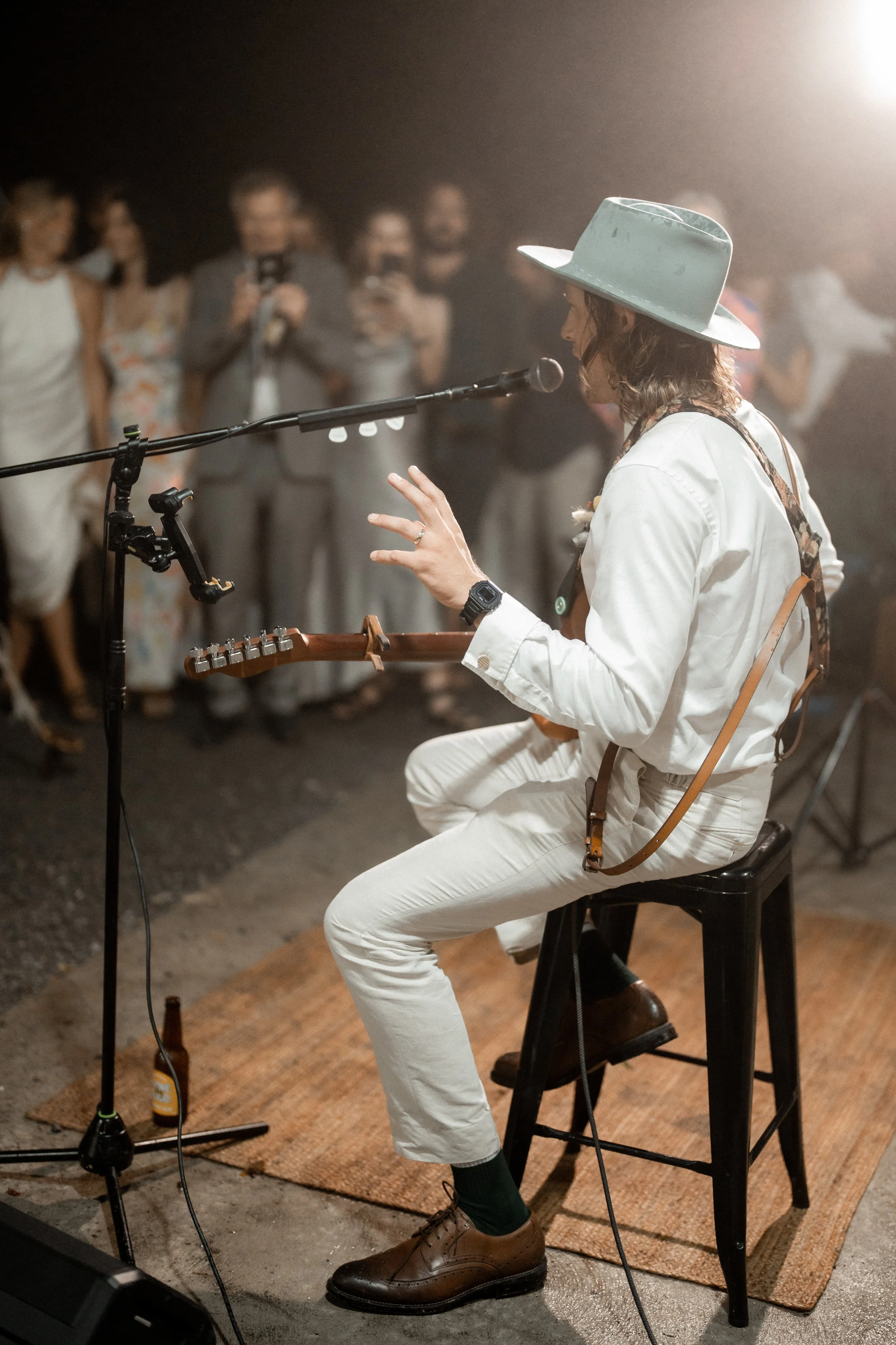 A musician in white clothes and a wide-brimmed hat sitting on a black stool, playing guitar and singing into a microphone on a stage, with an audience in the background.