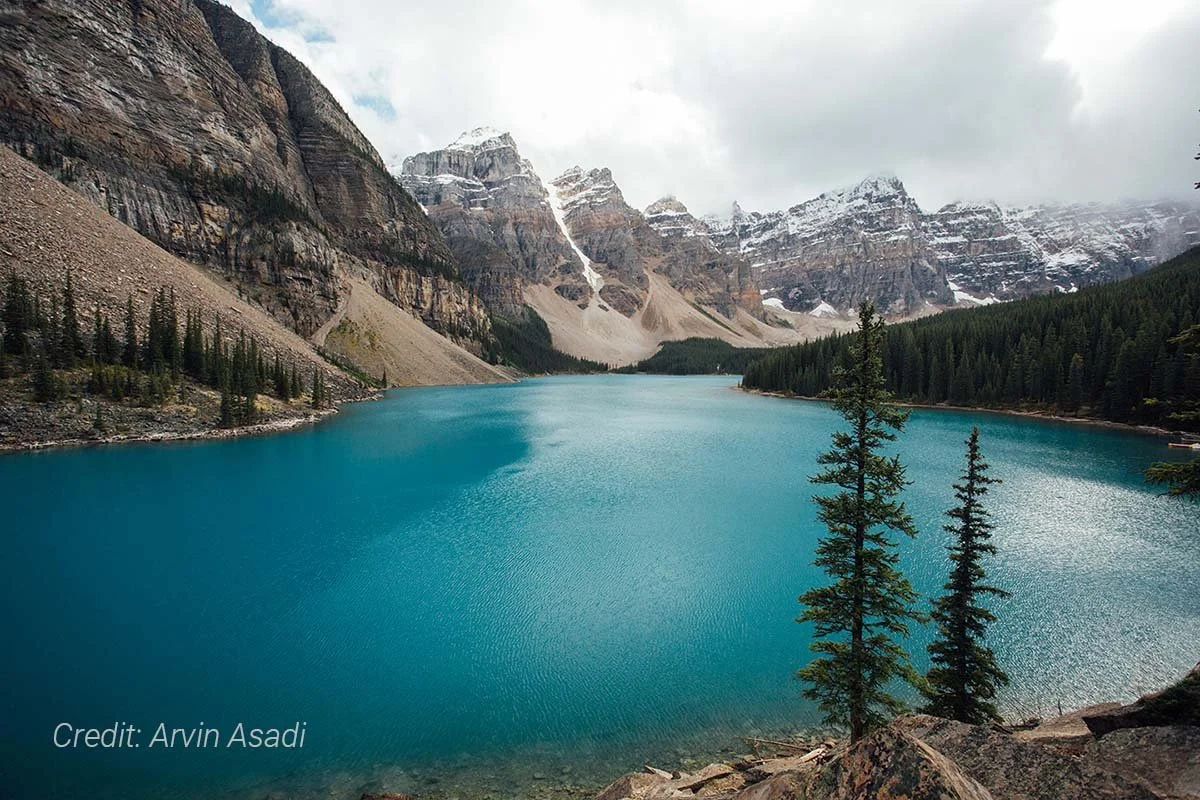 A scenic view of a turquoise lake surrounded by tall evergreen trees and rocky mountains with snow on their peaks, under a cloudy sky.