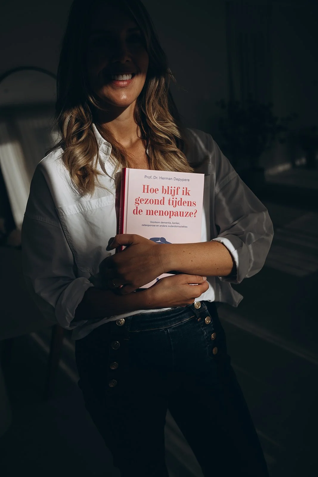 Delia Canini die glimlachend een boek vasthoudt met de titel 'Hoe blijf ik gezond tijdens de menopause?' in een donkere kamer.