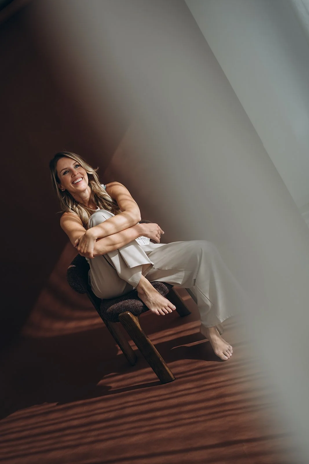 Een Delia Canini zit op een lounge stoel, gekleed in beige kleding, omringd door zacht licht en schaduw.