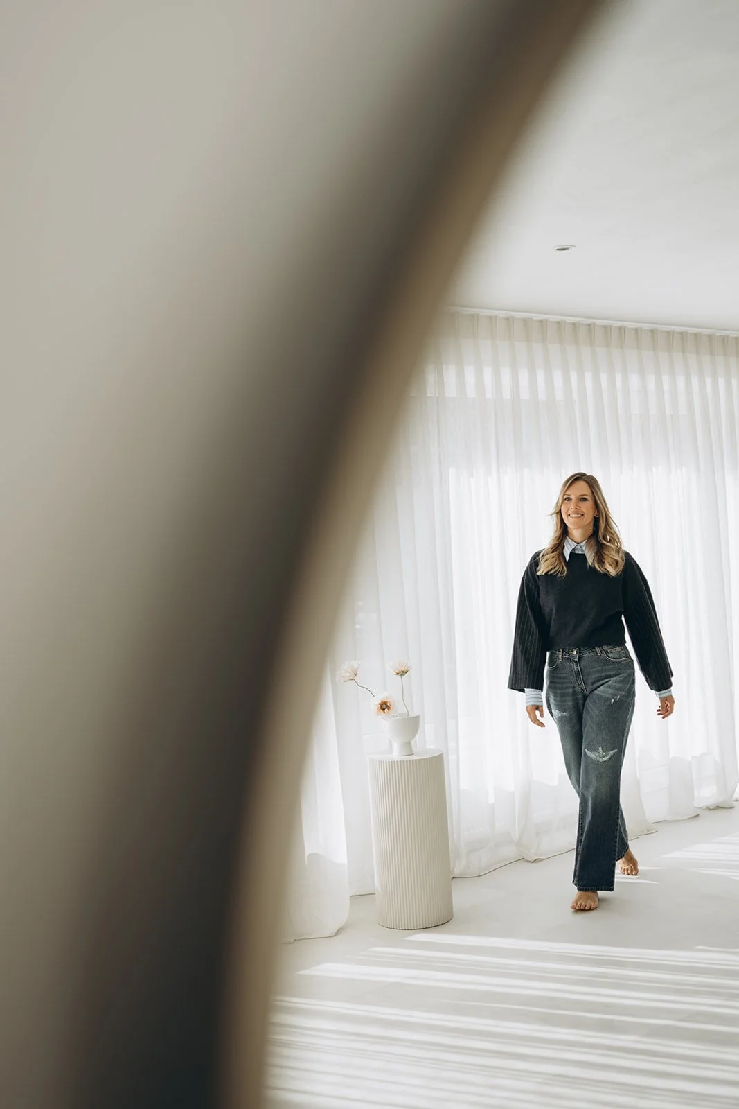 Delia, menopauze consulente die glimlachend in een lichte kamer staat met witte gordijnen en een vaas met bloemen, gezien door een uitsparing.