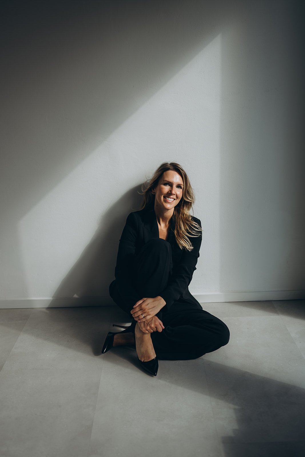 Delia Canini in zwart pak zittend op de vloer, lachend, in een ruimte met witte muur en lichte vloer, met schaduw van een raam op de muur.