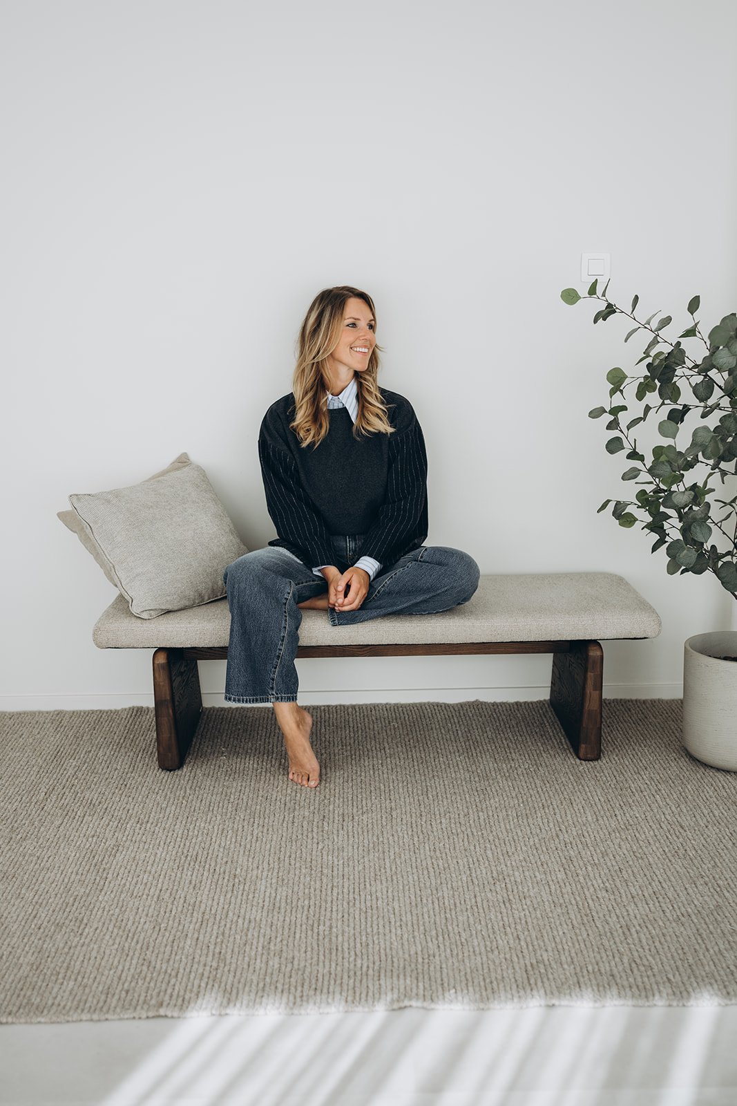 Delia, menopauze consulente zit op een bank in een minimalistisch interieur, glimlachend terwijl ze naar links kijkt. Ze draagt een donker trui en een lichtblauw blouse, met los haar.