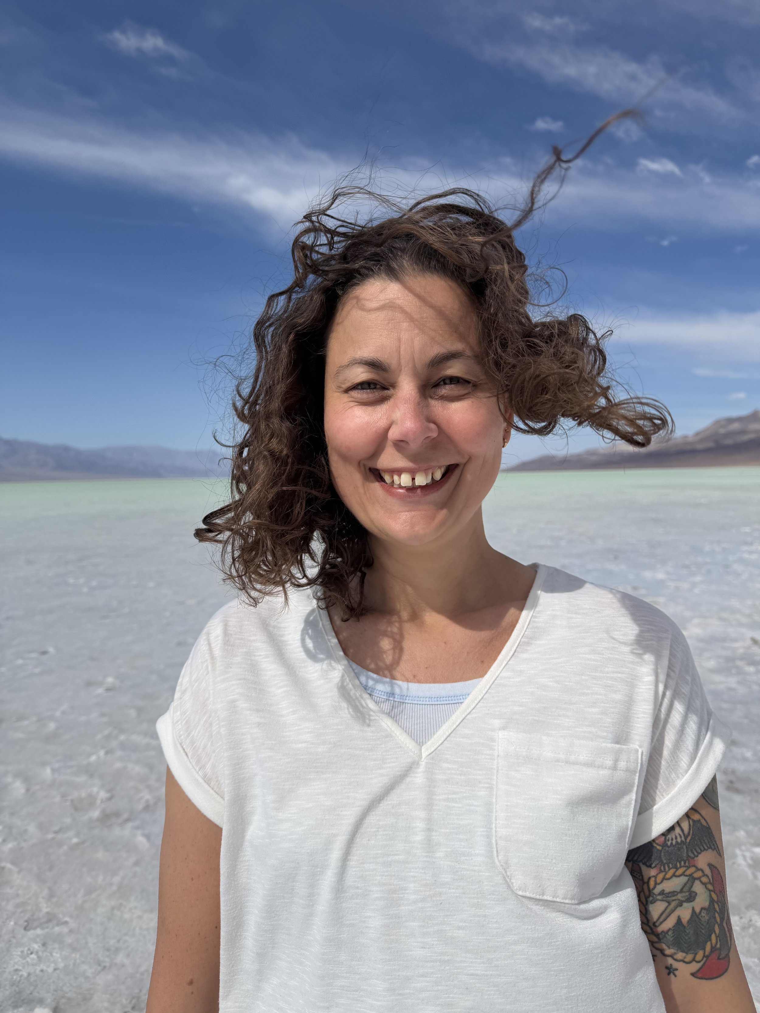 A woman healed from childhood trauma with curly hair smiling in a white T-shirt outdoors, with a mountain and blue sky in the background.