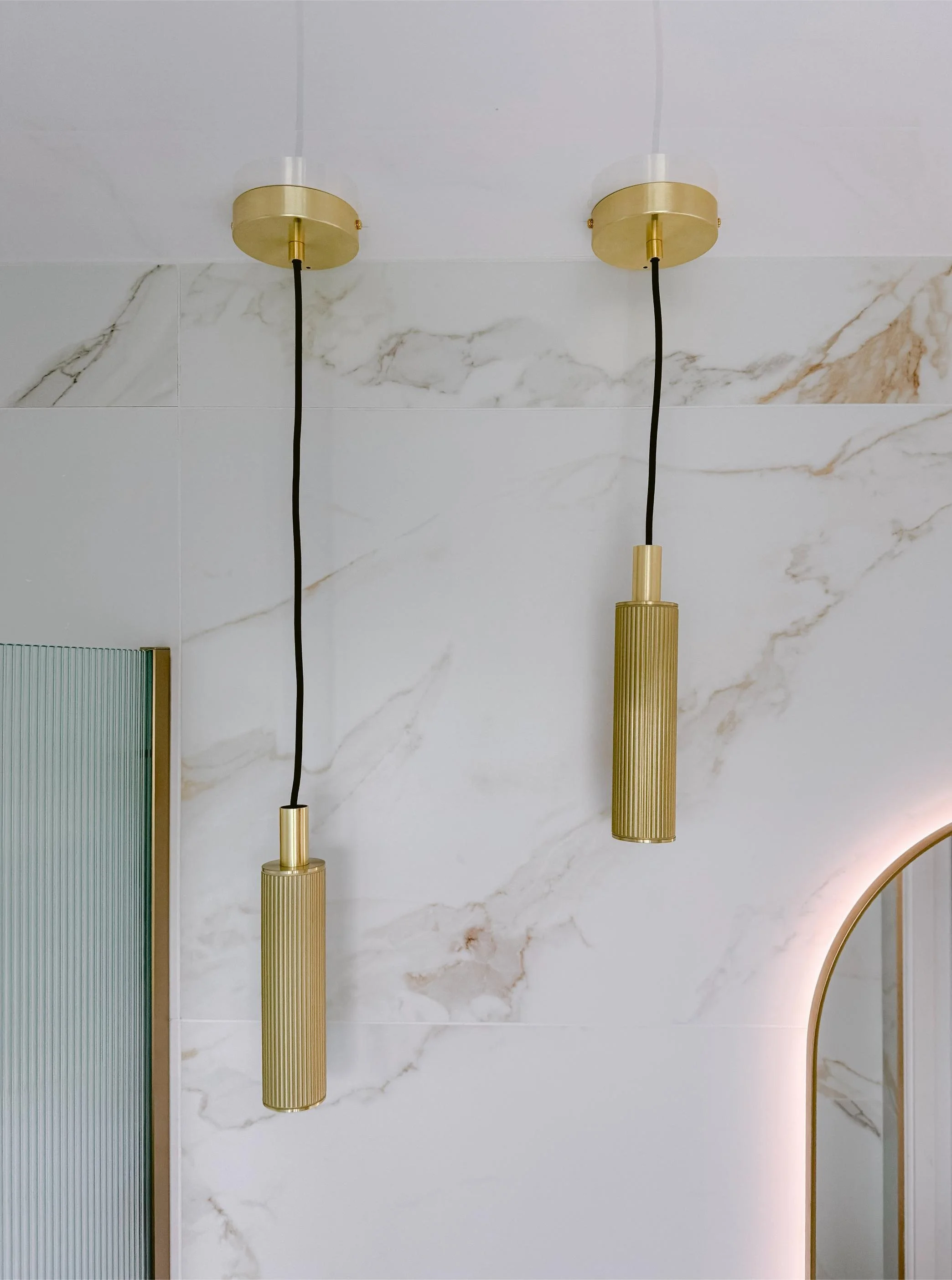 Two modern gold pendant lights with cylindrical ribbed designs hanging from the ceiling against a white marble wall.
