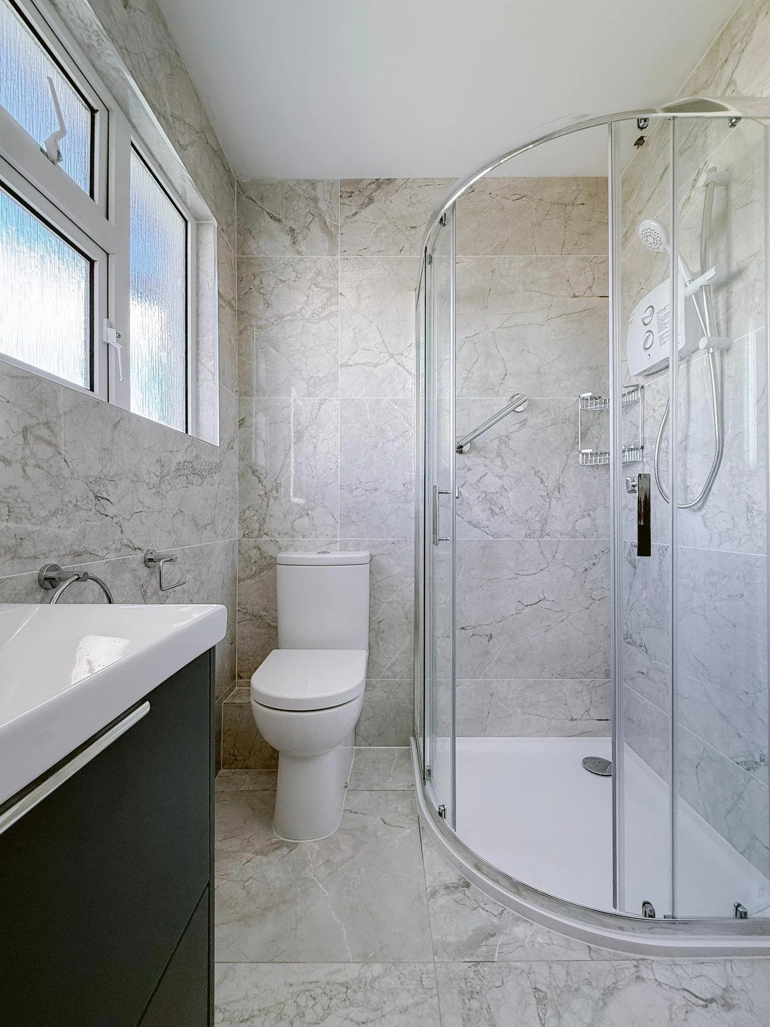 A modern bathroom with a frosted window, a white toilet, a black vanity with a sink, and a corner shower with glass doors and tiled walls.