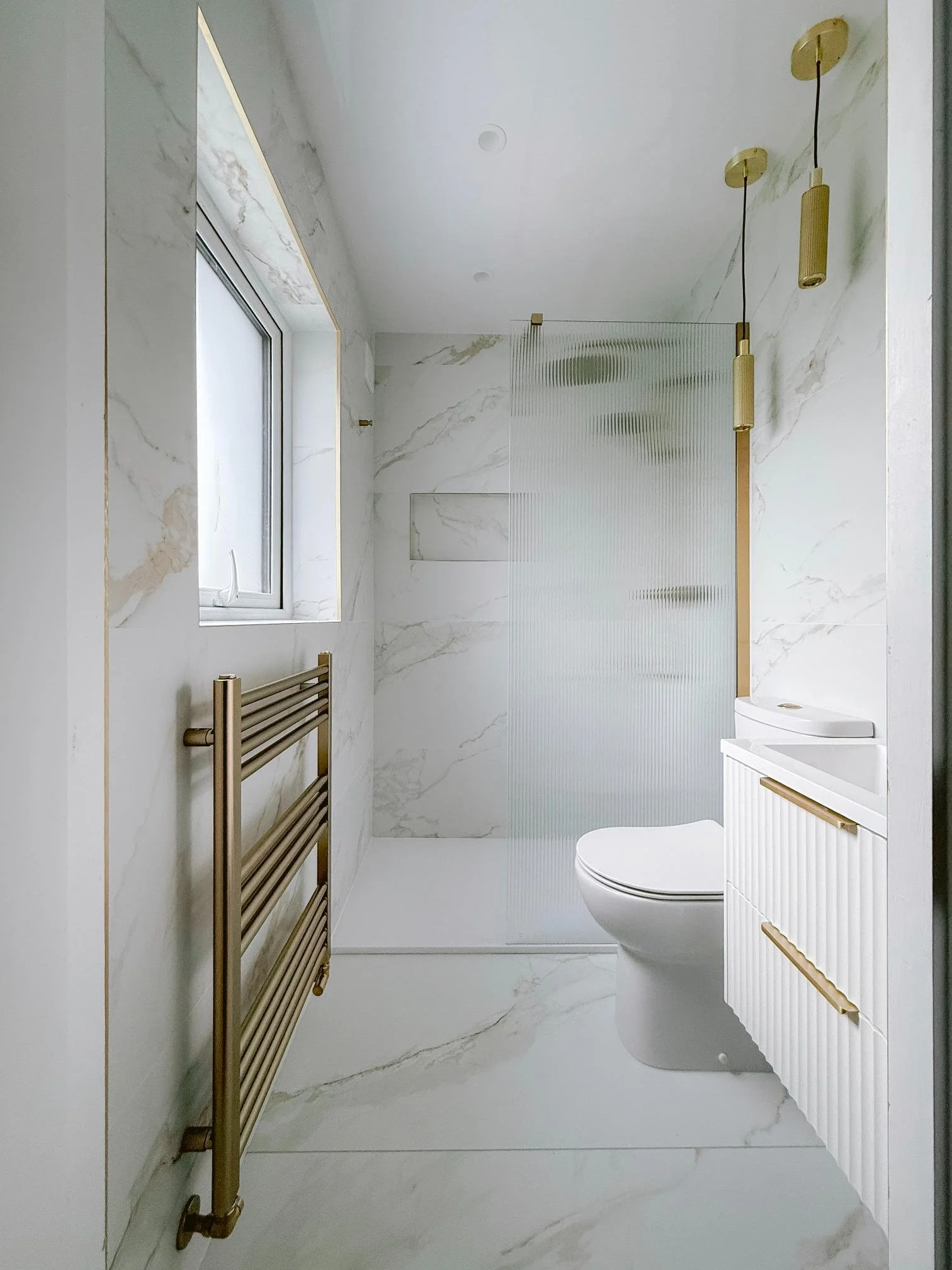 A modern bathroom with white marble walls and floor, a window, gold accents, a towel rack, a toilet, and a glass shower enclosure.