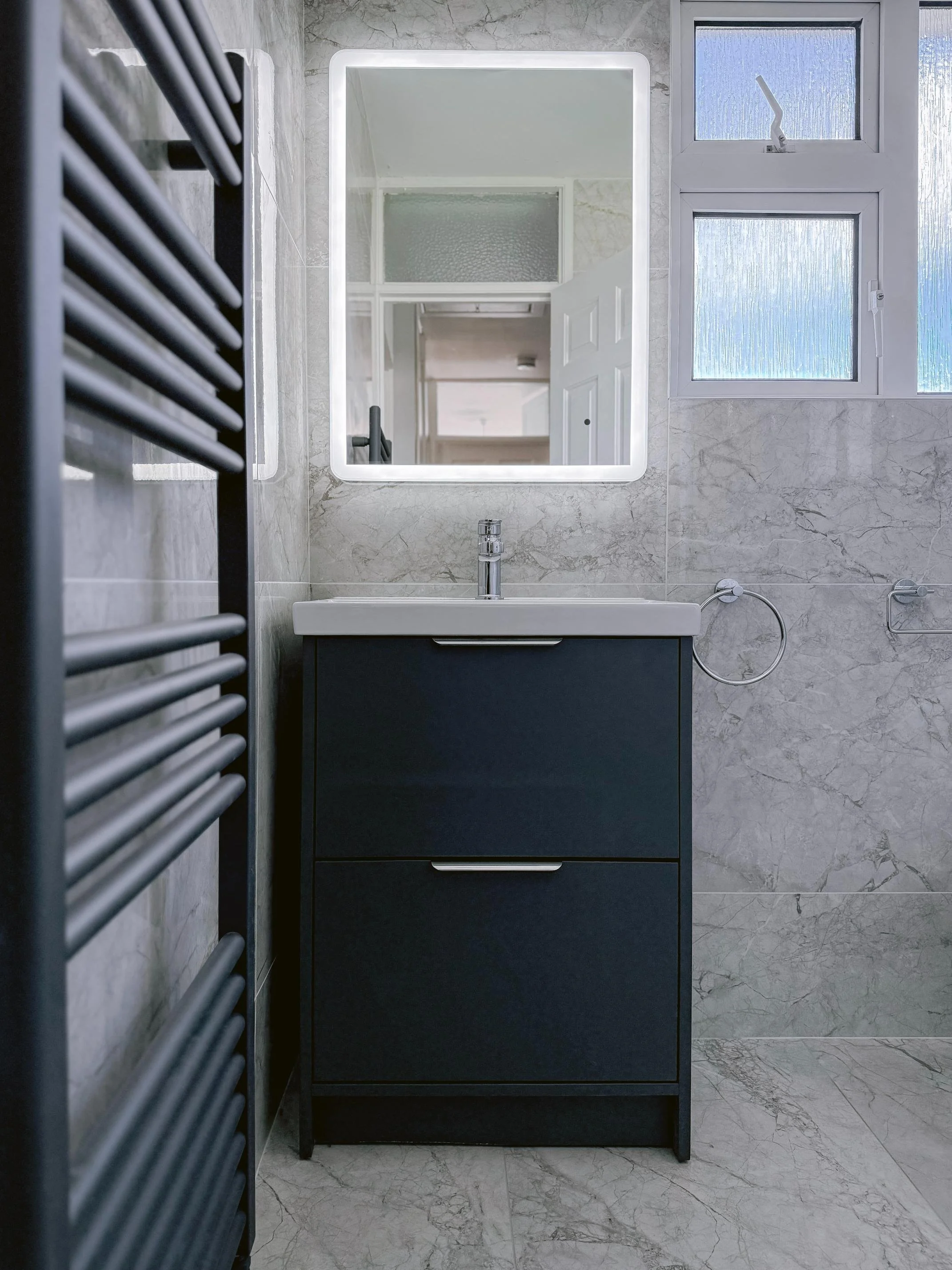 A modern bathroom featuring a rectangular mirror with LED lighting, a dark blue vanity with two drawers, a white rectangular sink, a chrome faucet, marble wall tiles, a window with frosted glass, and a black heated towel rail.