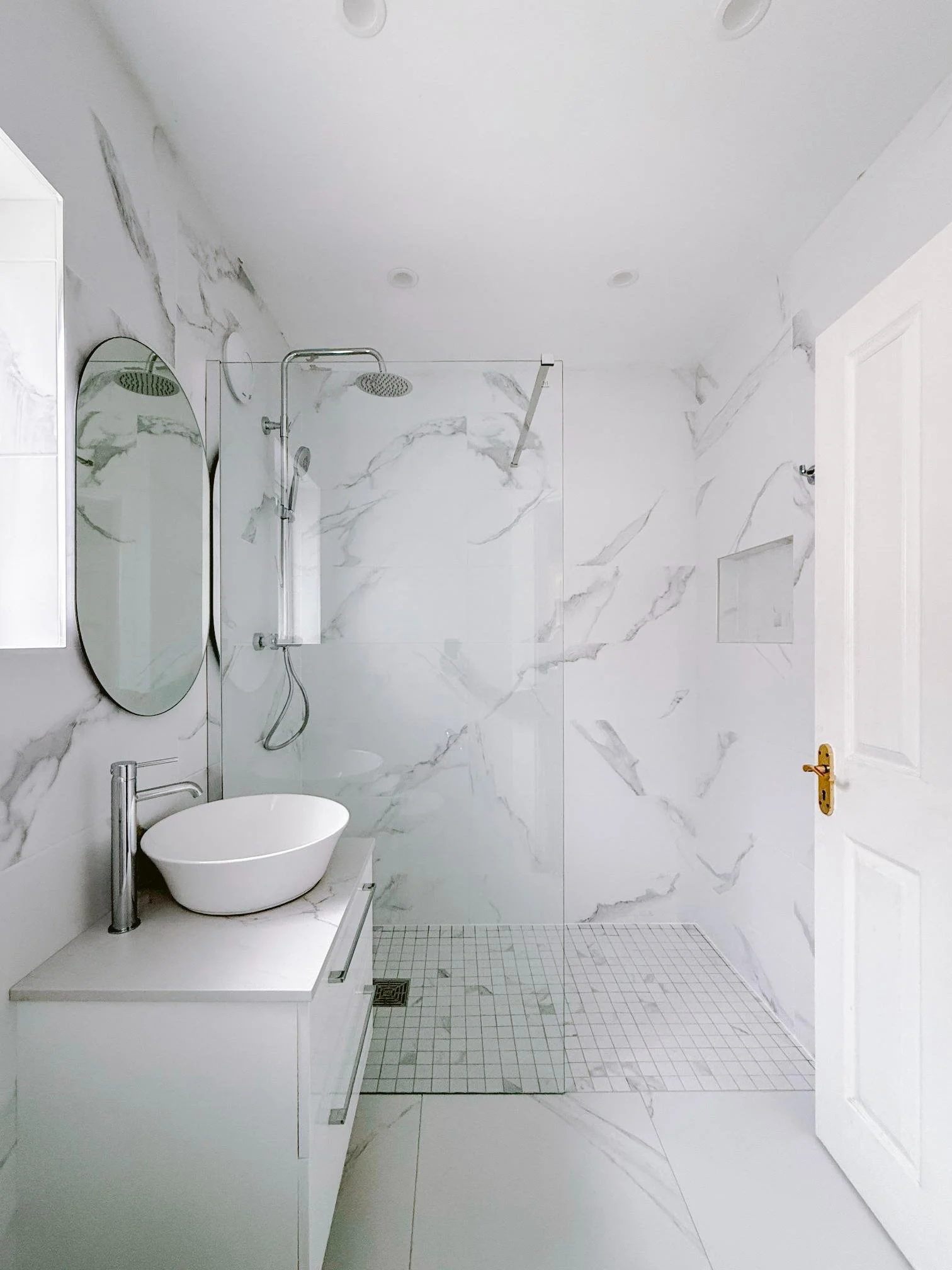 Minimalist bathroom with white marble walls, a walk-in shower with glass partition, a white vanity with a vessel sink, oval mirrors, and a white door, all illuminated by ceiling lights.