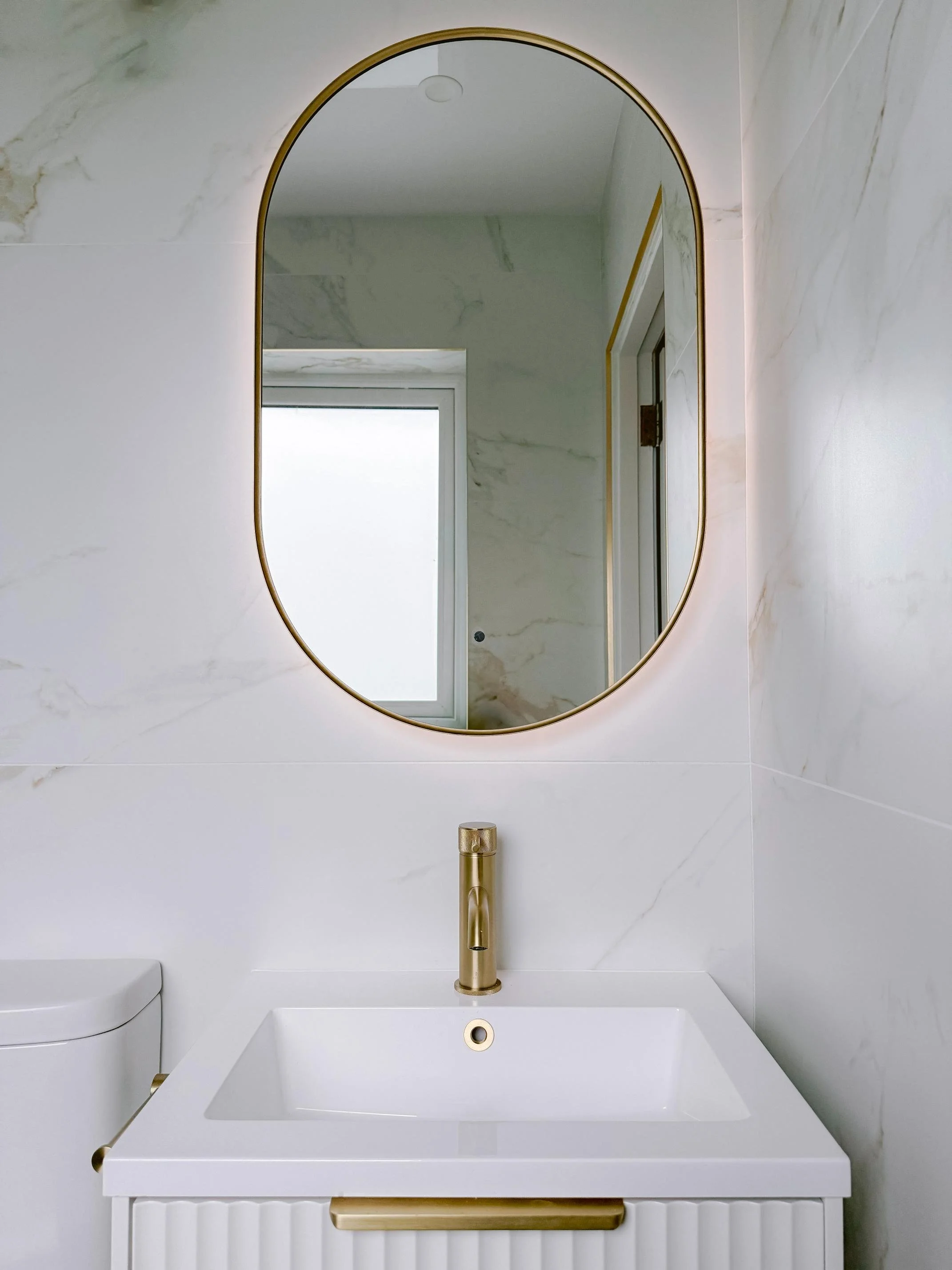 A modern bathroom sink with gold fixtures, a rectangular white basin, a gold faucet, a large oval mirror, a window with frosted glass, and marble-patterned wall tiles.