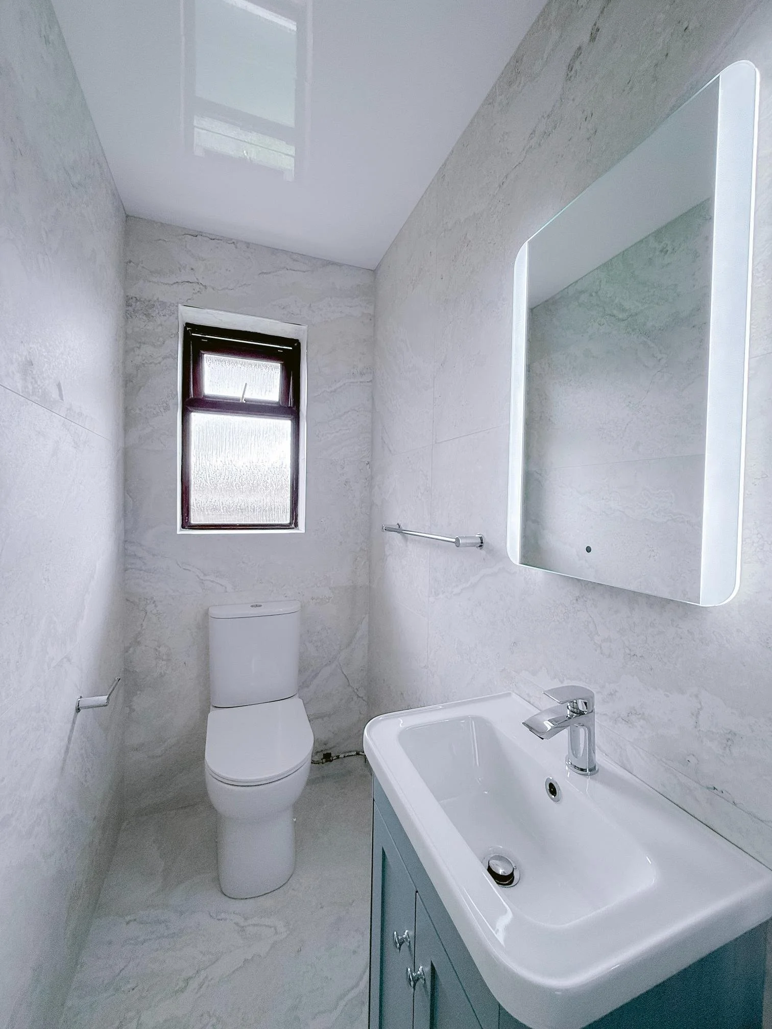 Small, modern bathroom with a white toilet, a white sink with a blue cabinet, a mirror with backlighting, a small window, and marble walls.