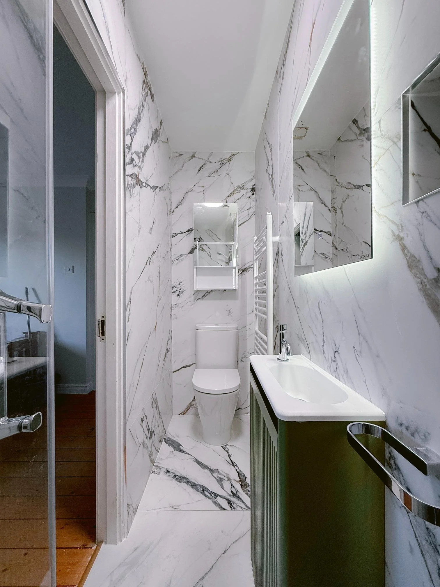 Modern bathroom with marble walls and floor, a toilet, a white sink with a green cabinet, a mirror, and a towel radiator.