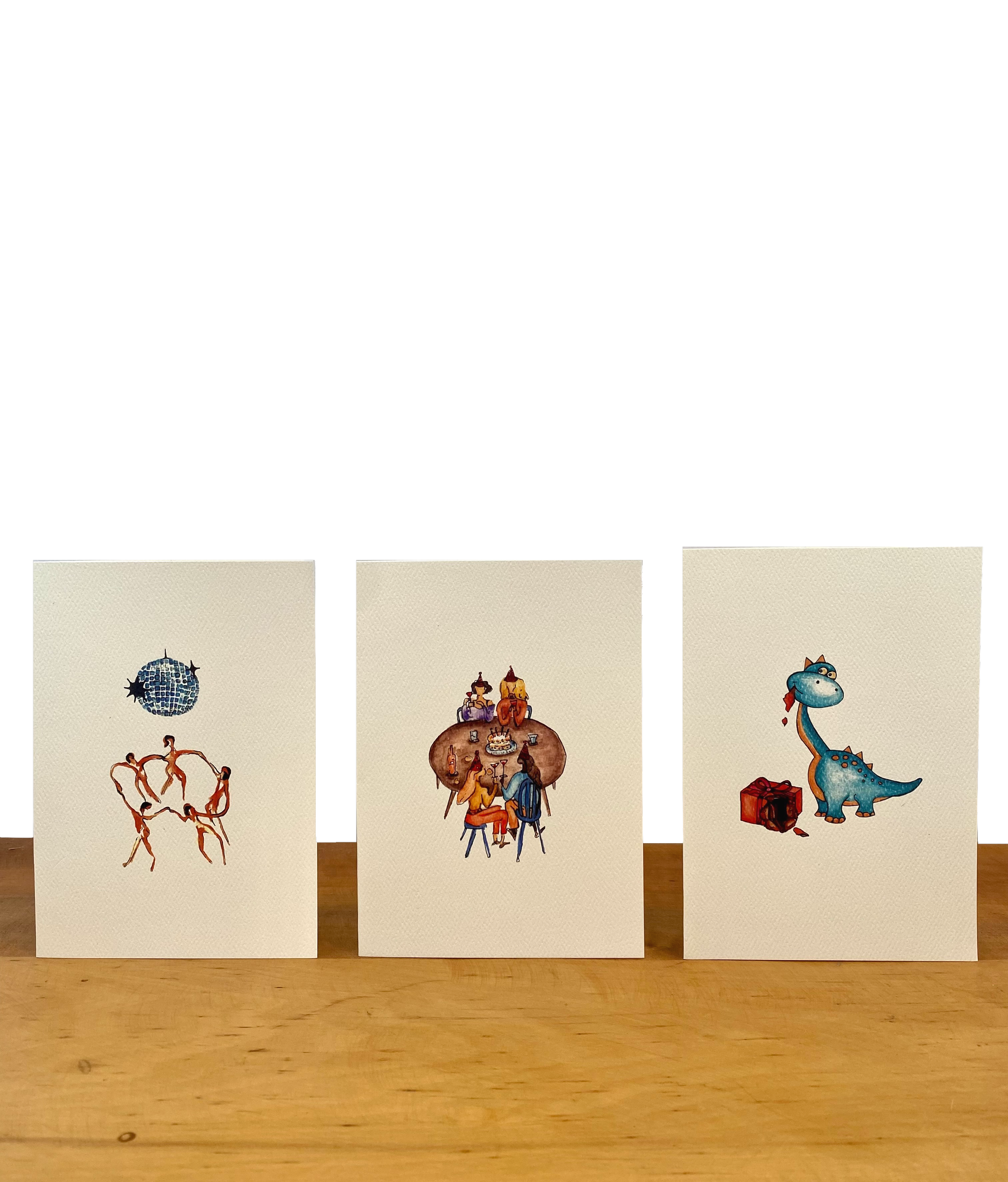 Three illustrated cards on a wooden surface. The left card features a disco ball and a circle of dancers. The center card shows a group of people sitting around a table enjoying a meal. The right card depicts a cartoon dinosaur with a present beside it.