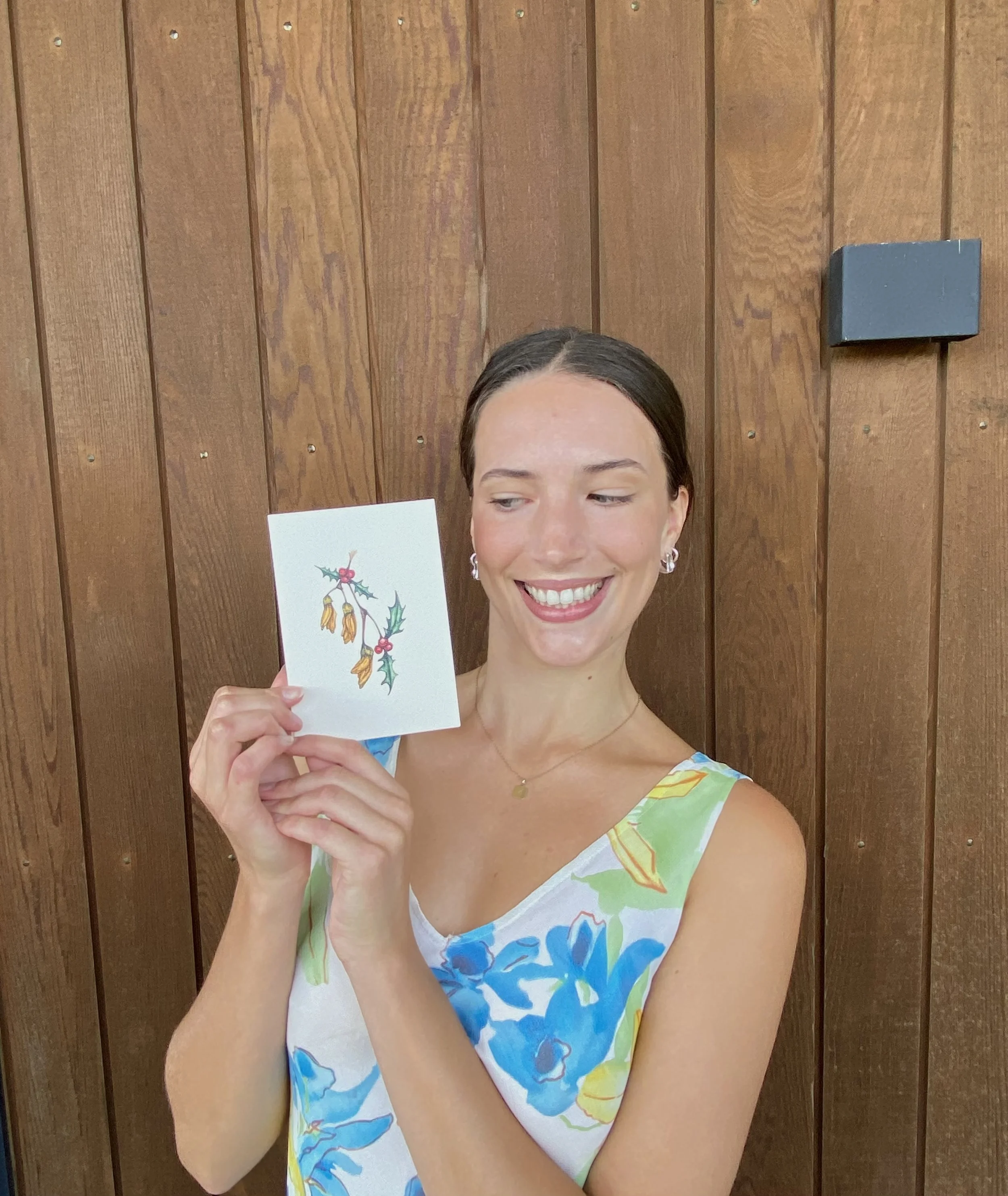 A woman smiling and holding a small card with a holiday-themed illustration of holly and kowhai, standing against a wooden wall.