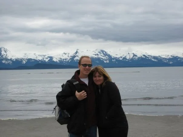 Laura Varnau, travel advisor, with husband standing by the water in Alaska with a snowy mountain range in the background