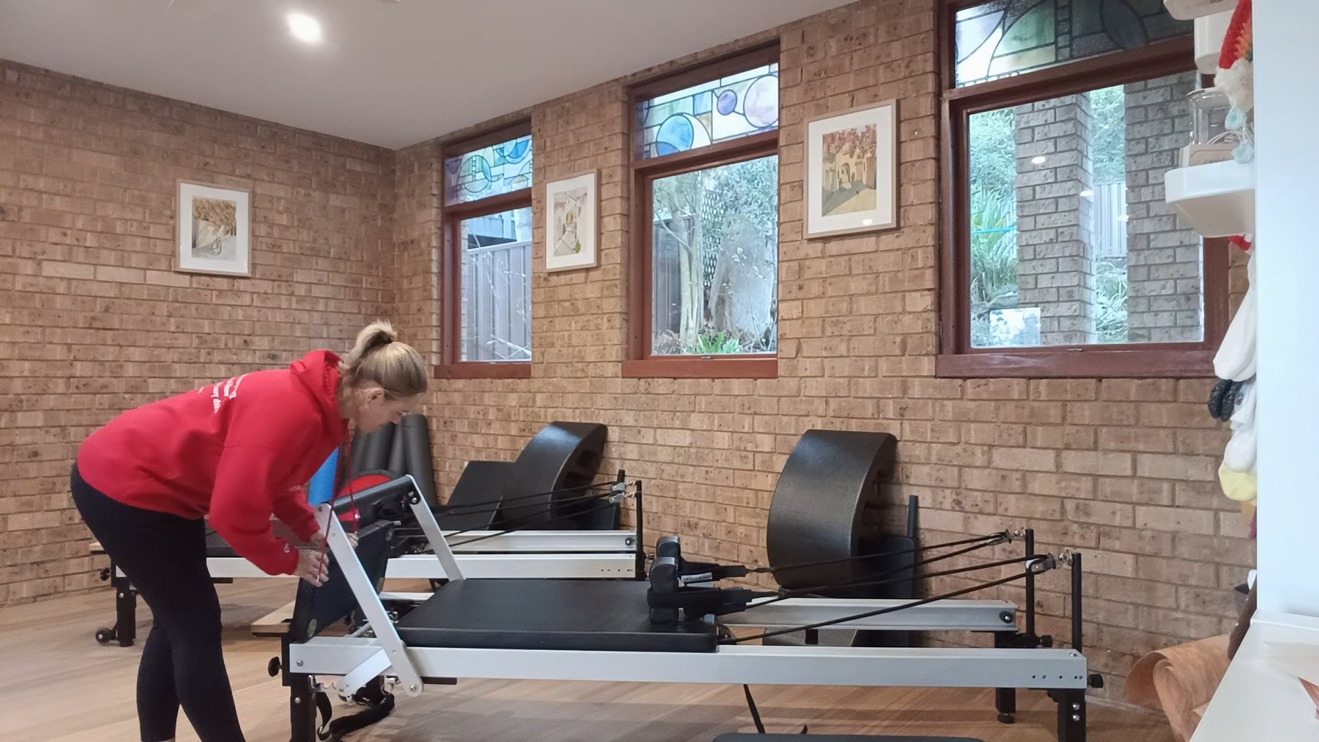 A woman in a red hoodie is adjusting or assembling a Pilates reformer machine in a room with brick walls and three large windows.