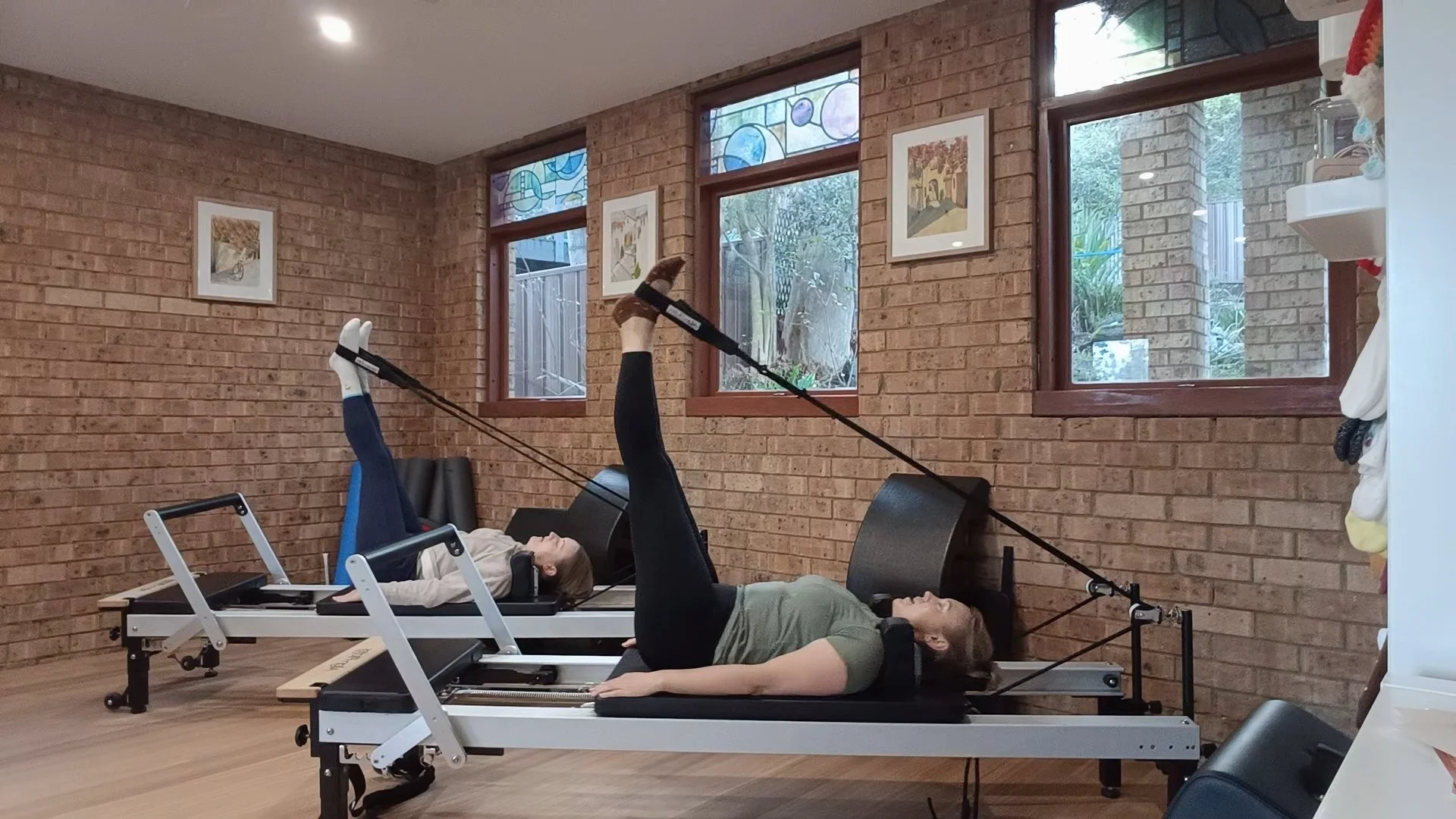 Two women practicing Pilates on reformer machines in a room with brick walls and large windows.