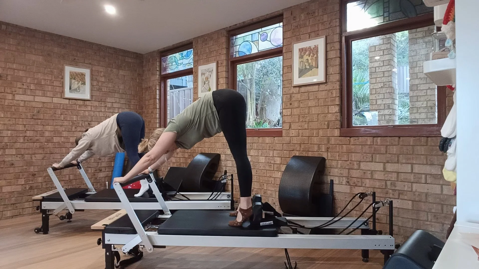 Two women are practicing Pilates on reformer machines in a sunlit room with brick walls, large windows, and framed artwork.