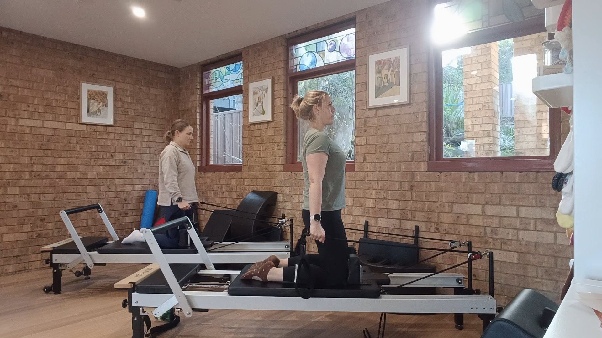Two women exercising on pilates reformer machines in a fitness studio with brick walls and windows.