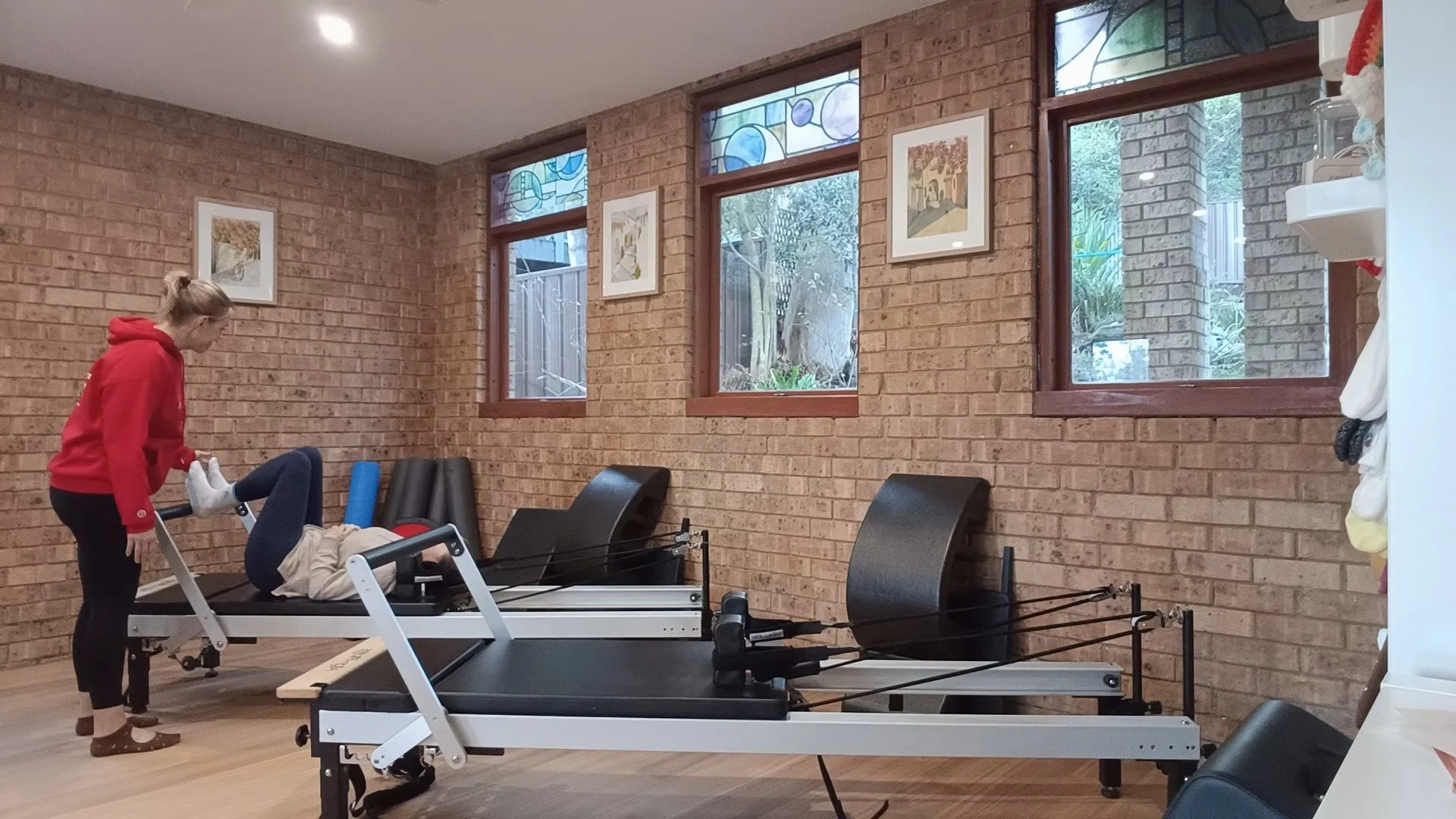A person assisting someone on a Pilates reformer machine in a brick-walled fitness studio with three large windows and some framed artwork.