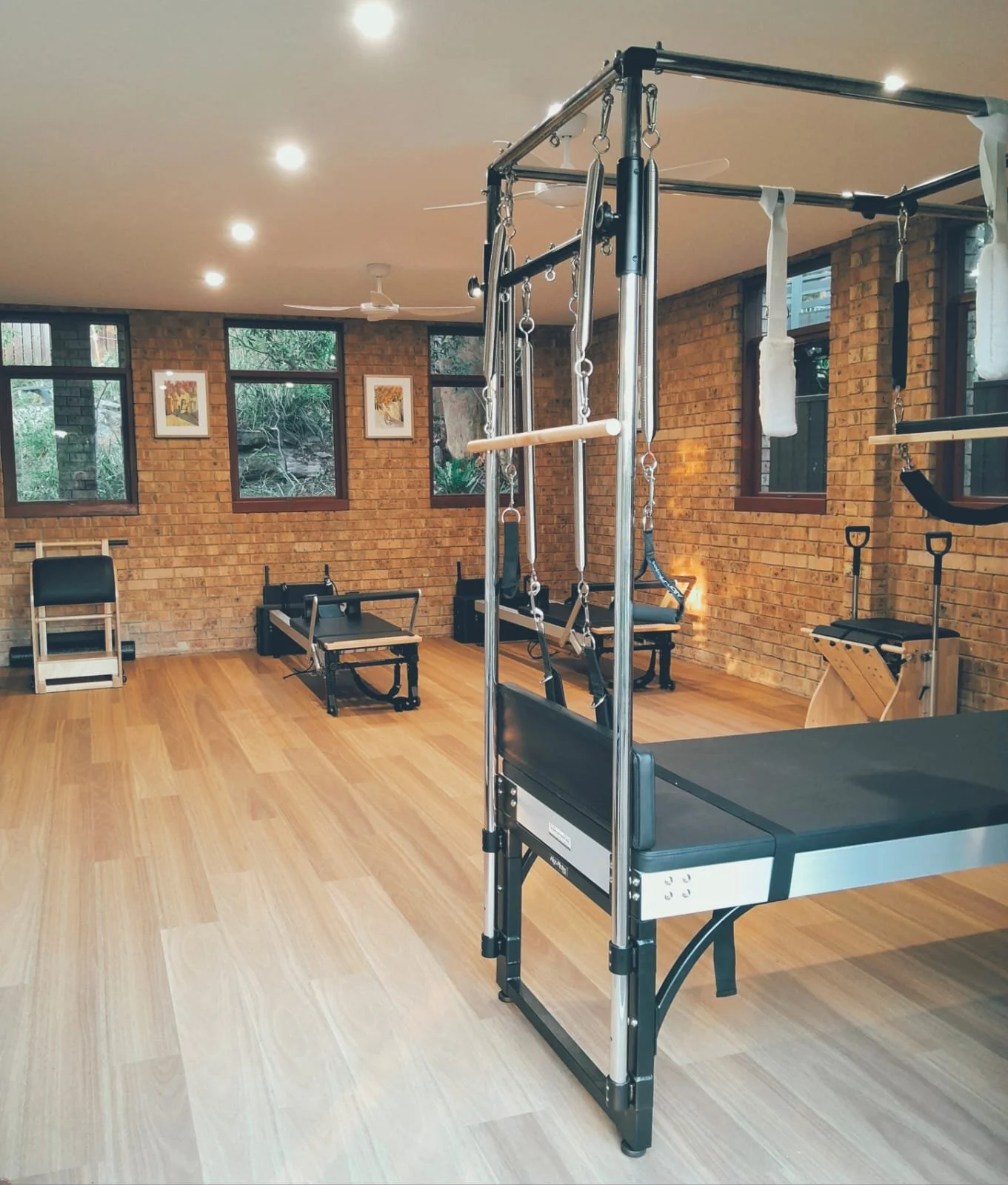A Pilates or physical therapy studio with brick walls, wooden floors, and large windows. Equipment includes a reformer, a Cadillac, a chair, and other Pilates apparatus.
