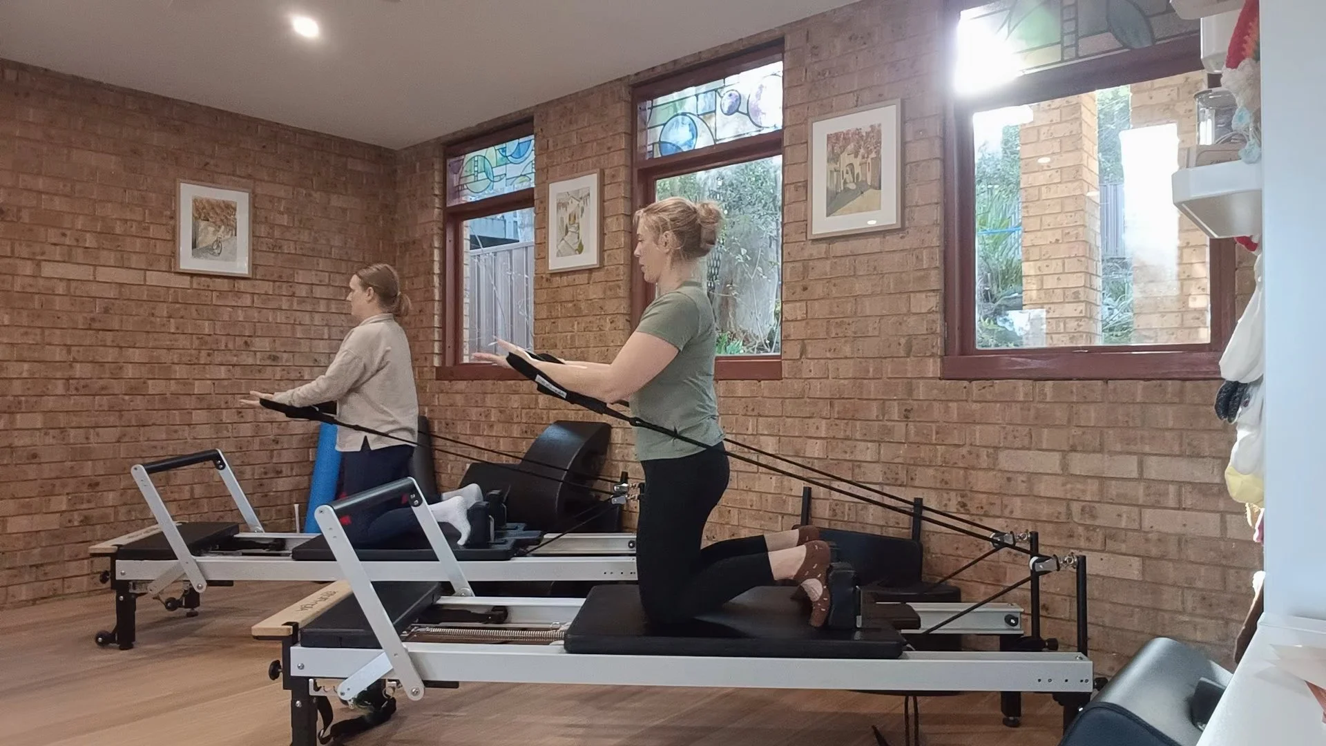 Two women exercising on reformer Pilates machines in a brick-walled fitness studio, with large windows and artwork on the wall.