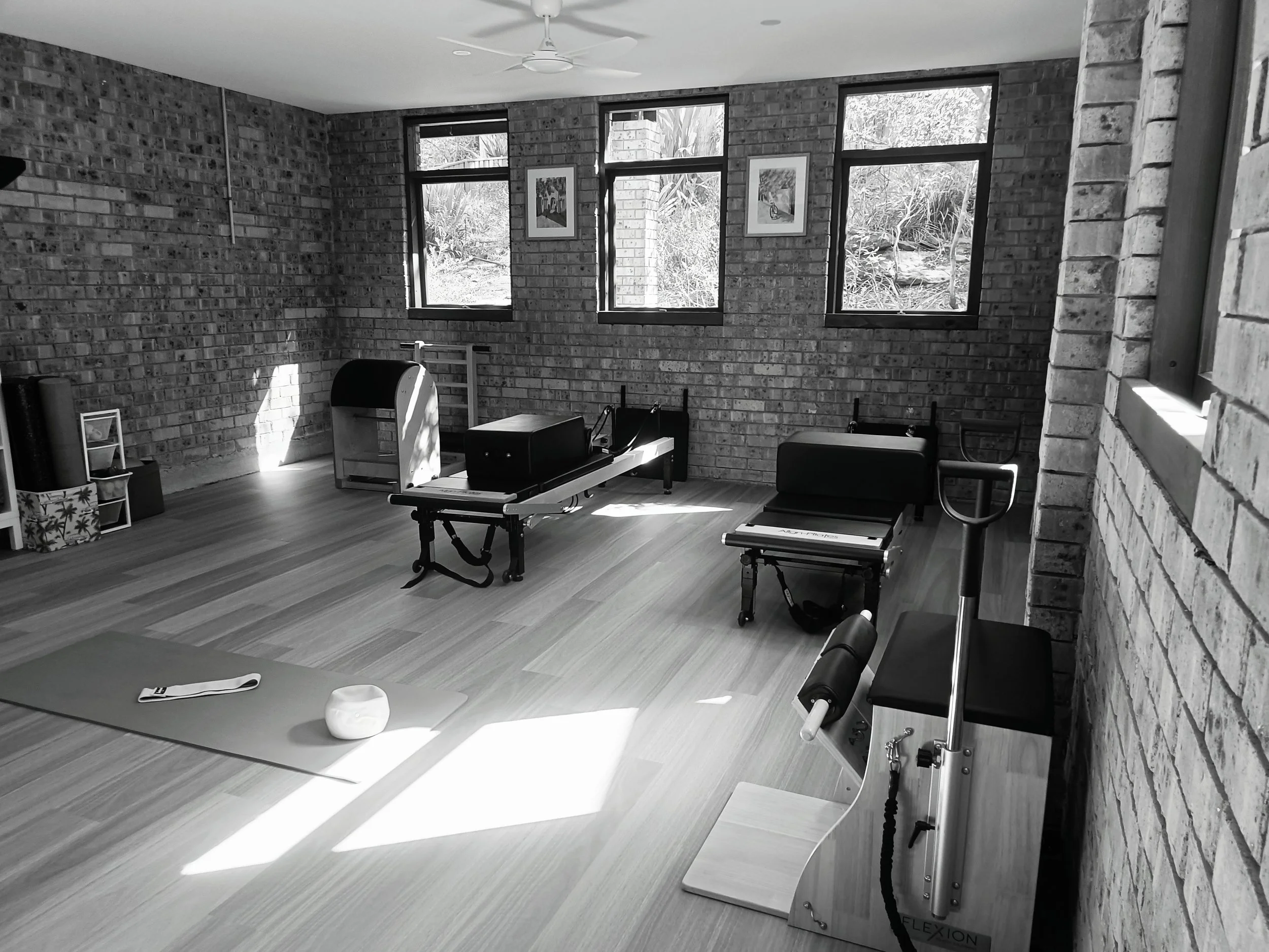 A therapy or Pilates room with large windows showing outdoor greenery. The room contains exercise equipment such as a reformer, a chair, and a barrel. The walls are brick, and there are framed pictures hanging. There's a ceiling fan in the room.