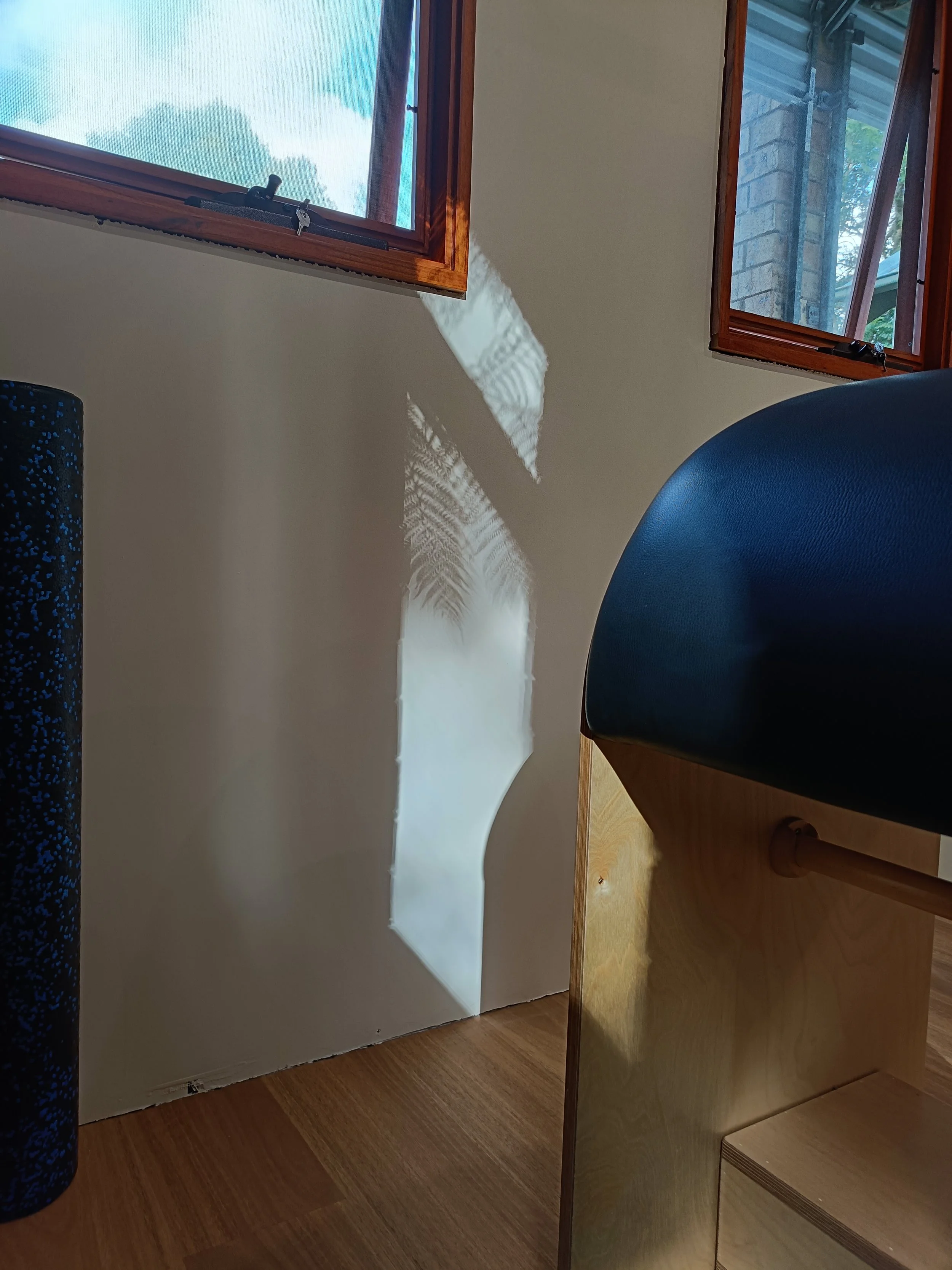 Open window with wooden frame, sunlight creating shadow patterns on a beige wall, part of a Pilates ladder barrel visible.